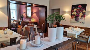 A dining area with tables set for two, featuring white tablecloths, candles, and potted plants.