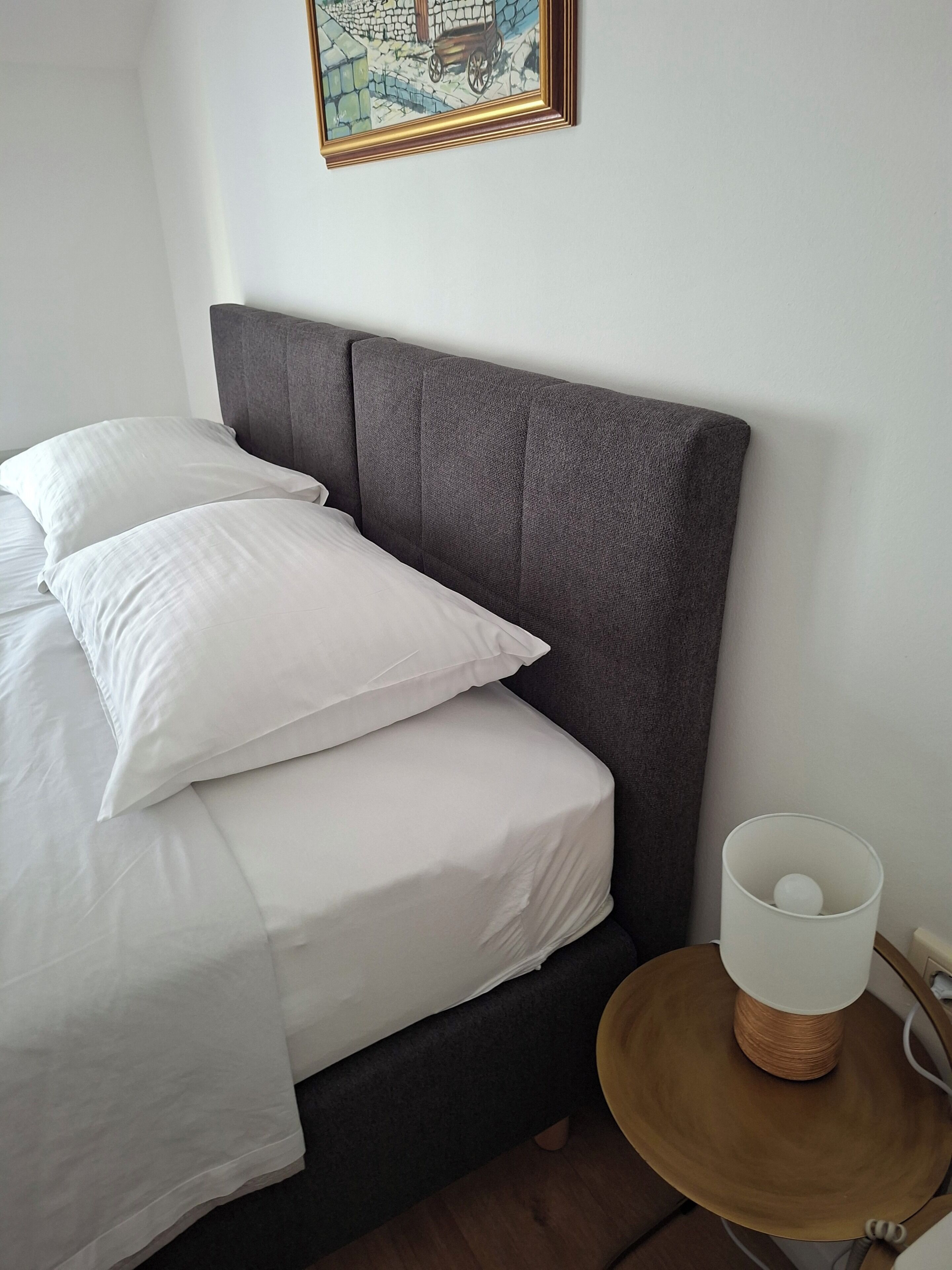 Una cama bien hecha con sábanas blancas, un cabecero tapizado en gris y una mesita de noche con lámpara.