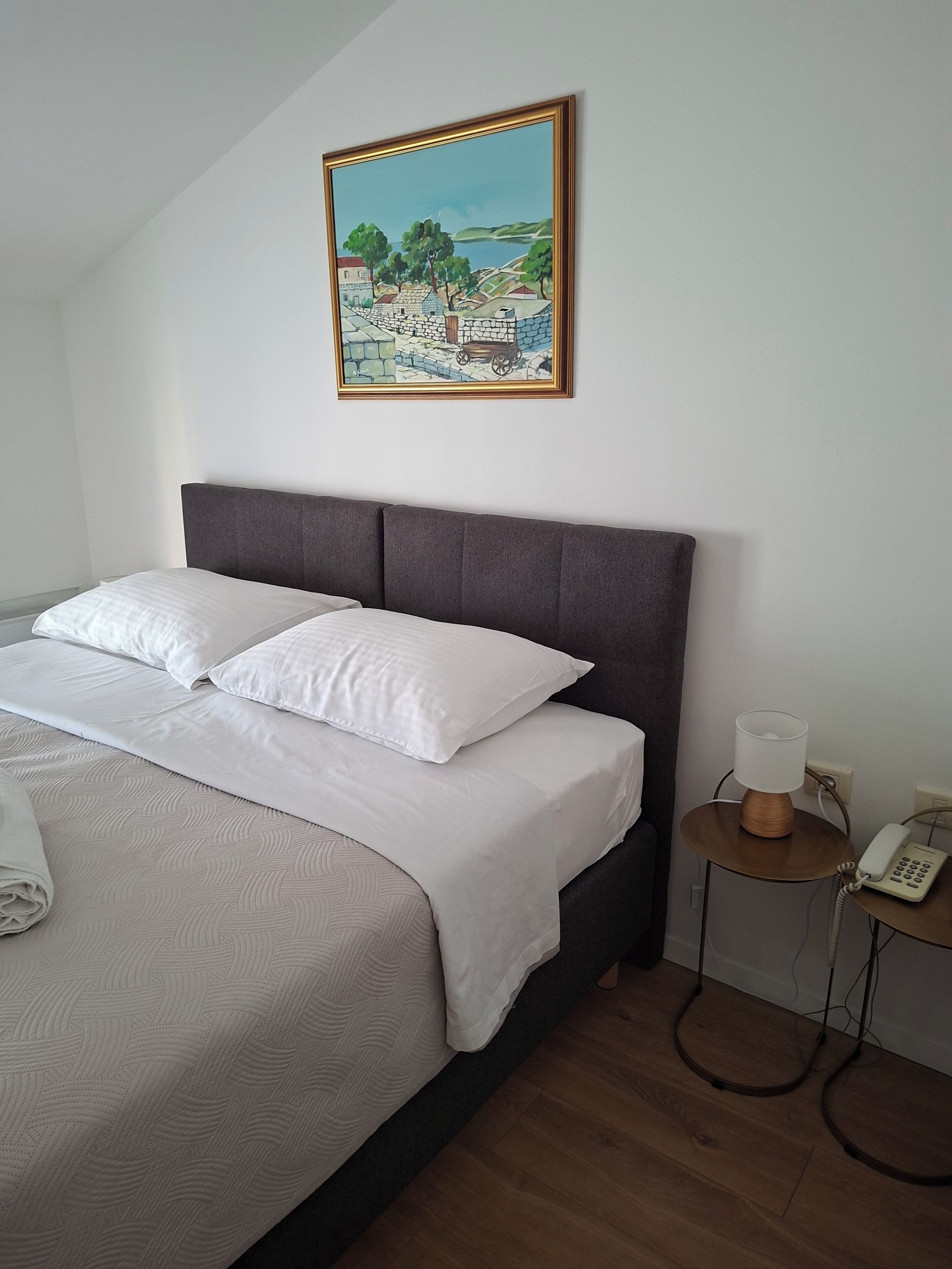 A neatly made bed with white linens, a grey upholstered headboard, and a painting of a landscape above it.