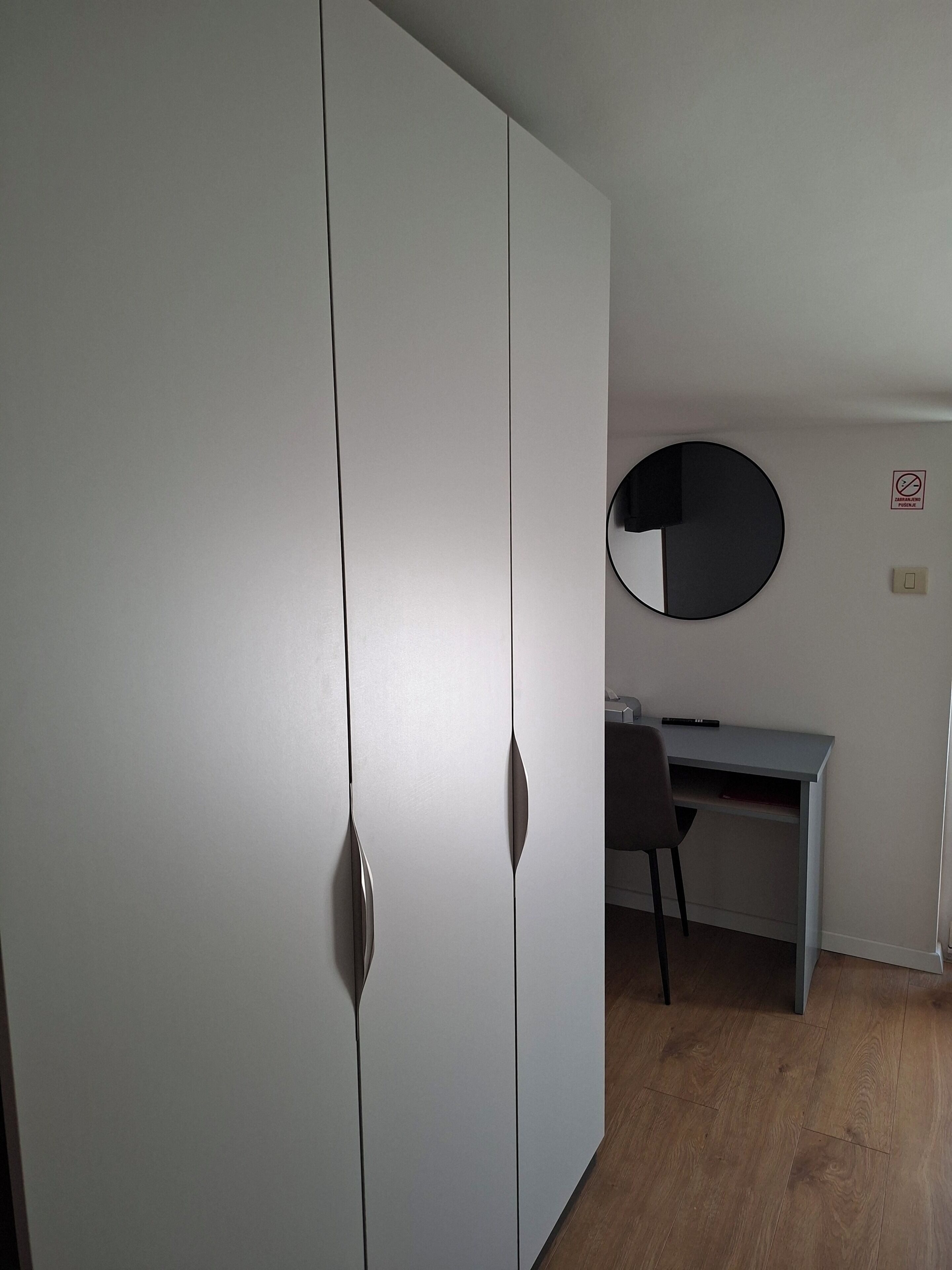 A room with a wardrobe, a desk, a chair, and a round mirror on the wall.