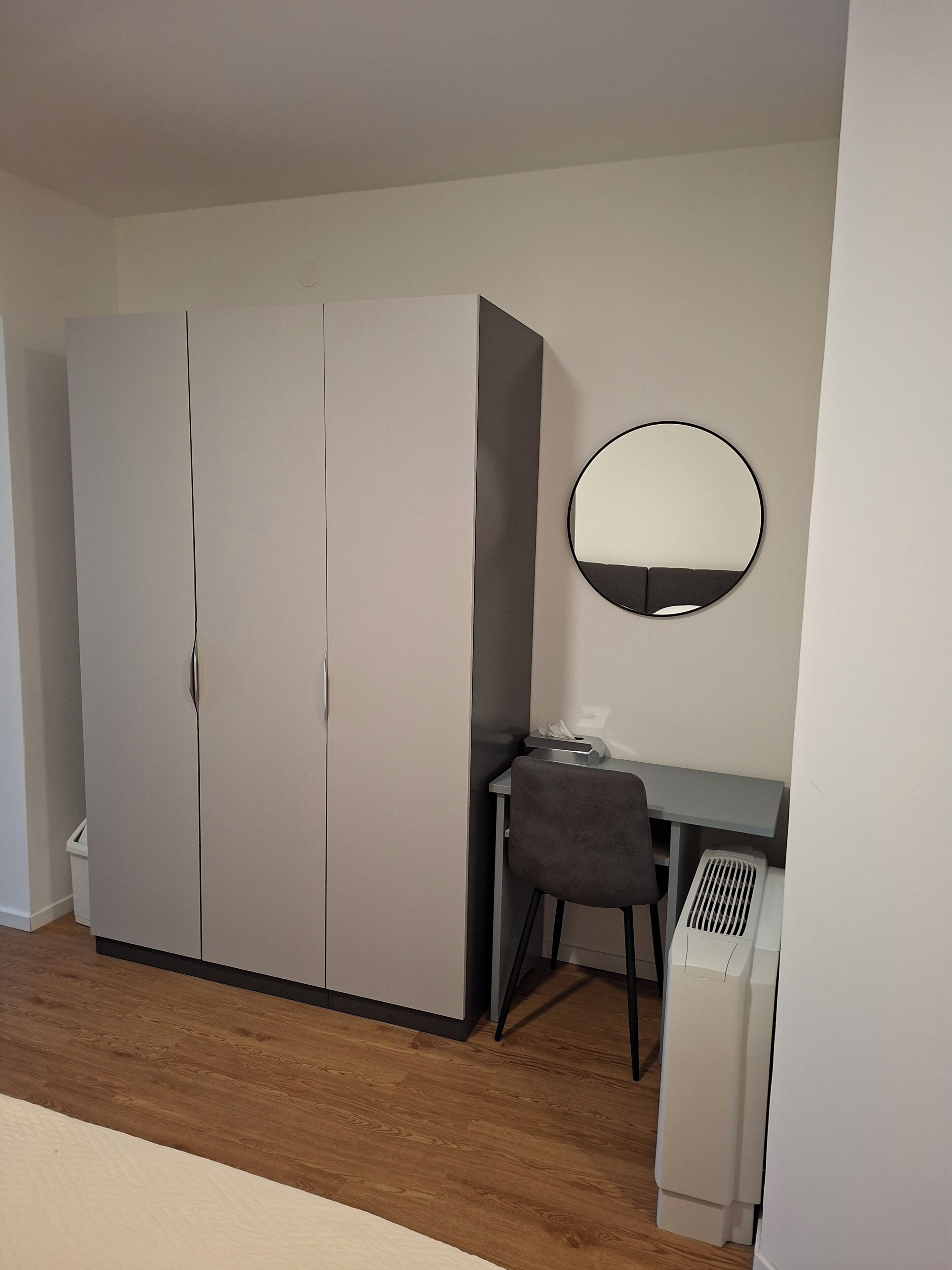 Una habitación con armario, escritorio, silla, espejo redondo y radiador blanco.