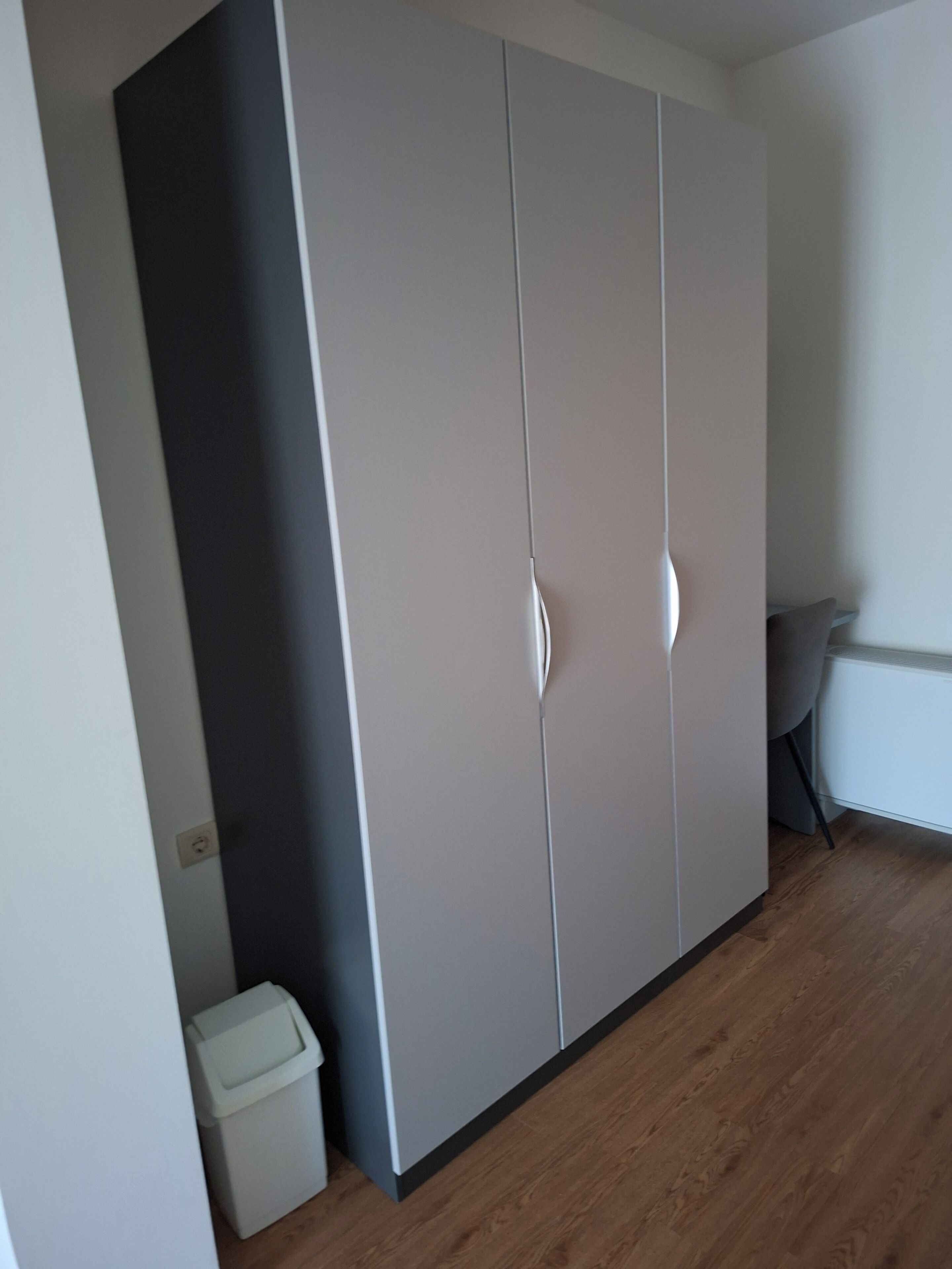 A room with a large wardrobe, a small trash bin, and a grey chair.