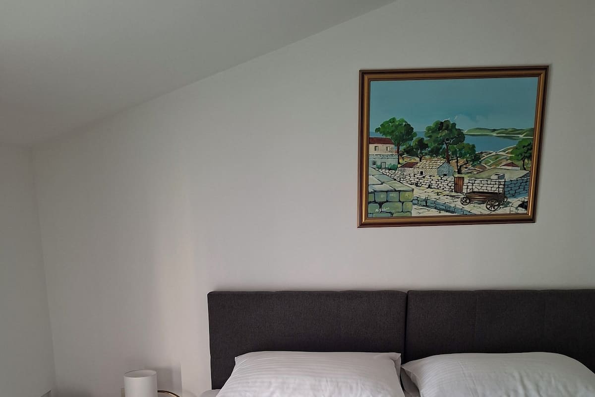 A neatly made bed with white linens, a painting of a landscape on the wall, and a small table with a lamp beside the bed.