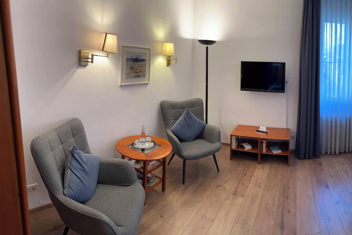 Une chambre d’hôtel avec un lit, deux fauteuils, une petite table, un téléviseur et une fenêtre avec des rideaux.