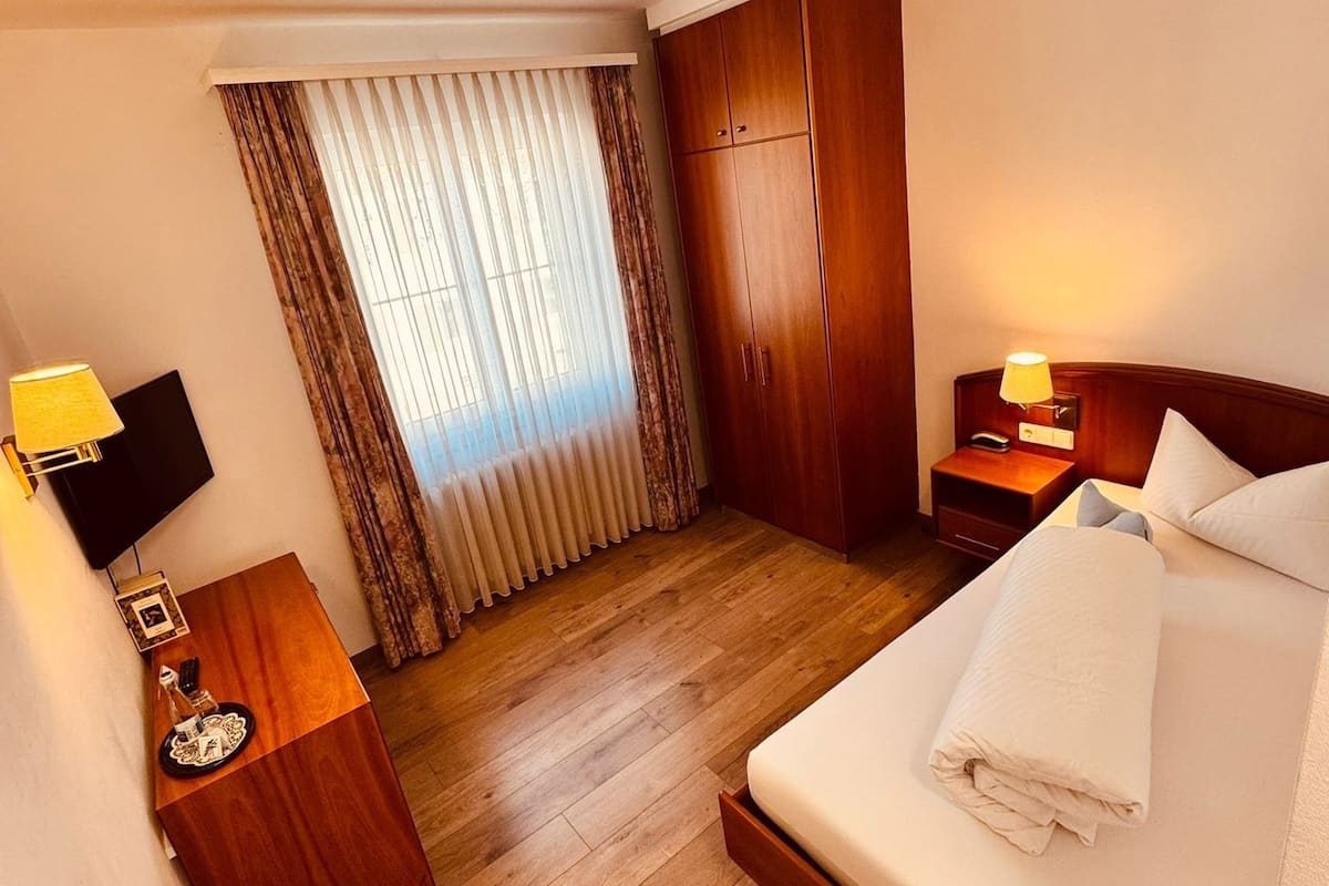 Une chambre d’hôtel comprenant un lit, une table de chevet, une lampe, une télévision, une armoire et une fenêtre avec des rideaux.