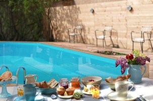 A poolside breakfast setup with a variety of food and drinks, including pastries, fruits, and a vase of flowers.