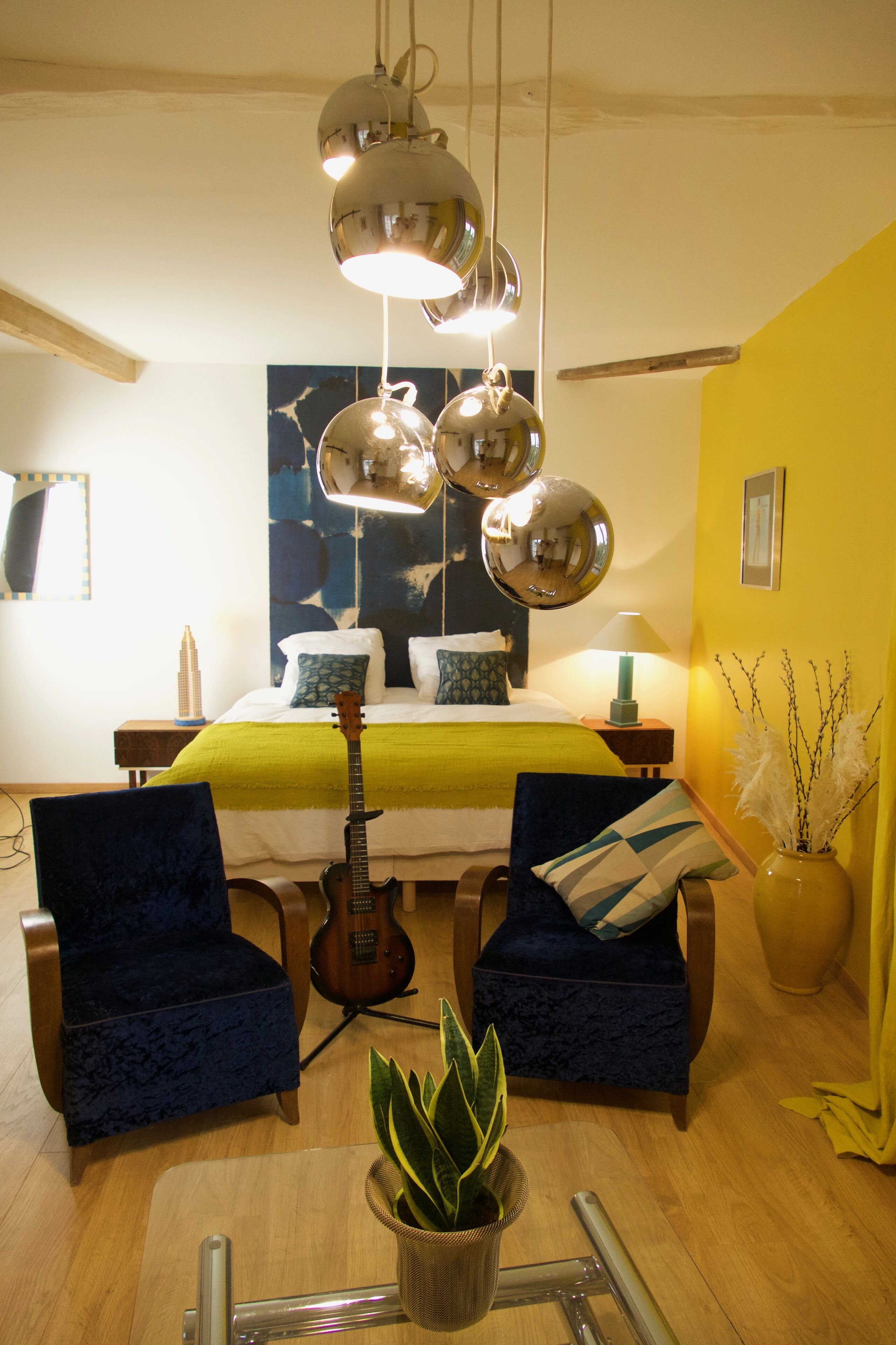 A room with a bed, two armchairs, a guitar, a plant, and a dining table with chairs.