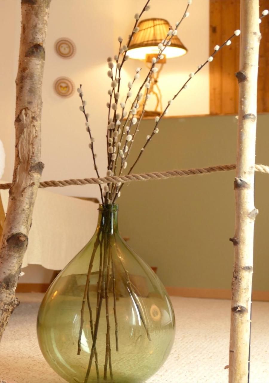 A clear glass vase with branches and small white flowers, placed on a surface with a rope and a lamp in the background.