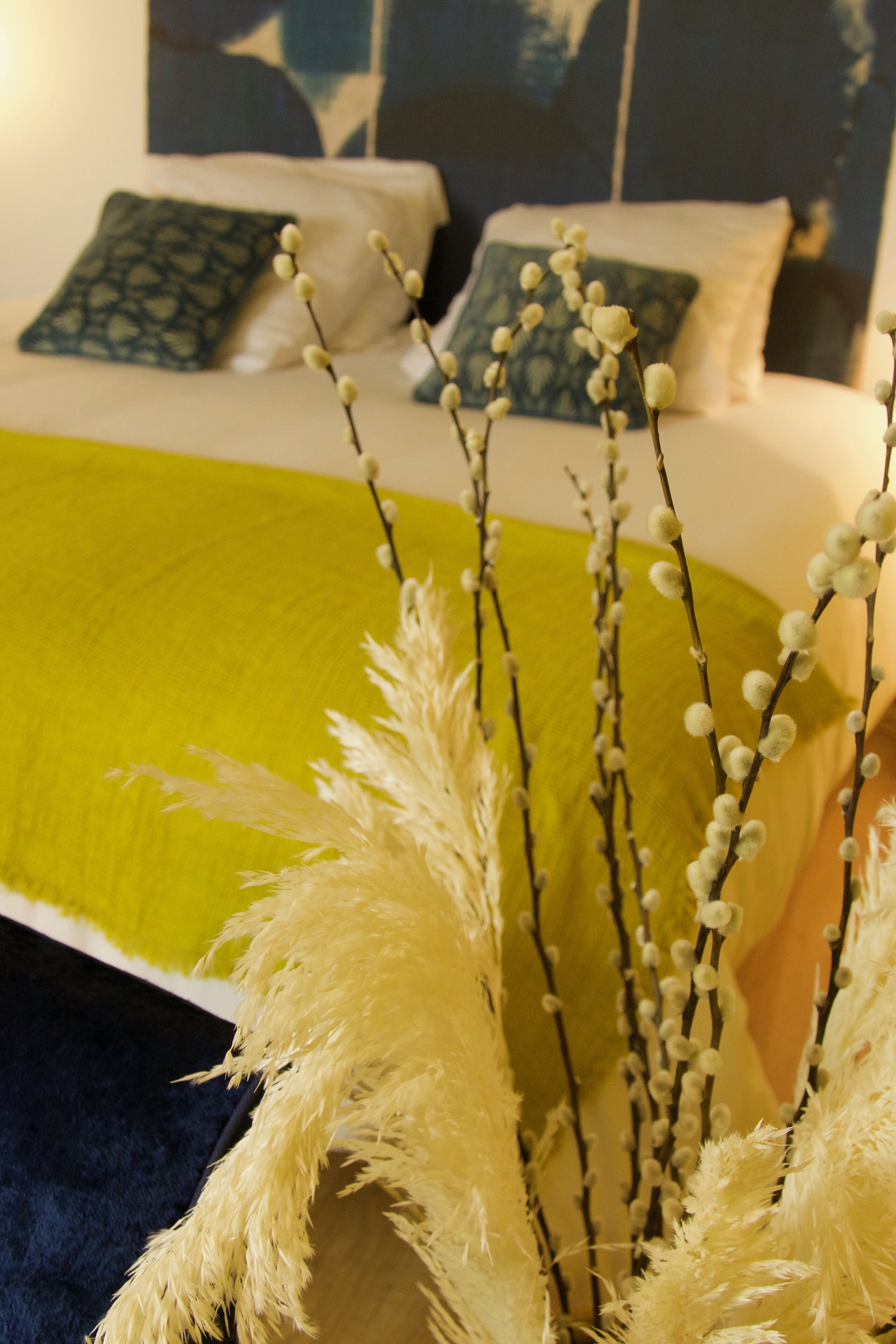 Una cama con un plaid verde, almohadas blancas y una rama decorativa con flores blancas.
