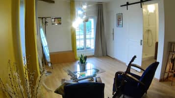 A spacious living room with a glass coffee table, a blue armchair, a guitar, and a potted plant.