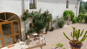 Un patio con un banco de madera, una mesa y sillas, rodeado de plantas en macetas y un cactus.