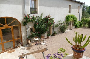 A courtyard with a wooden bench, a table, and chairs, surrounded by potted plants and a cactus.