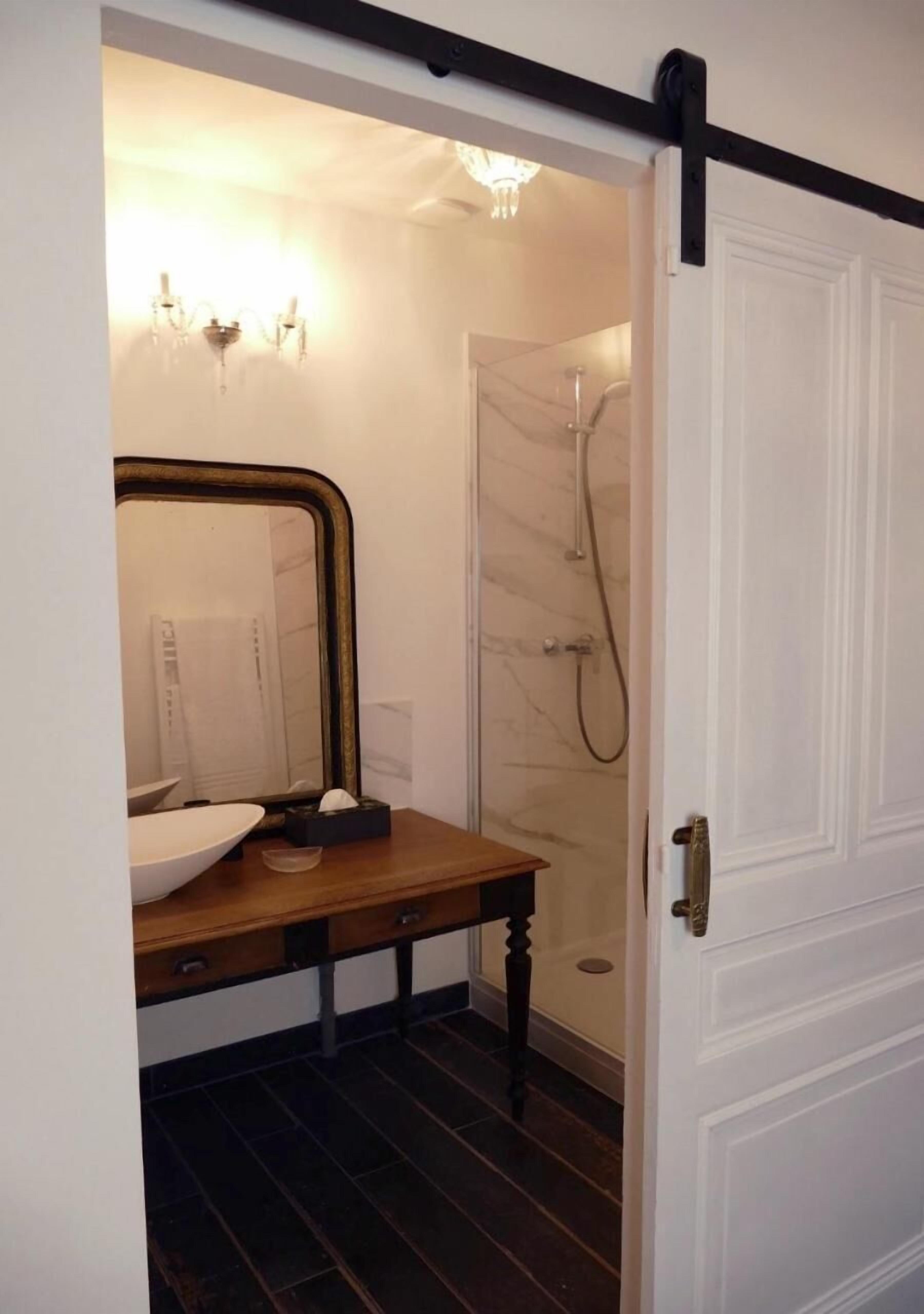 A bathroom with a wooden vanity, a large mirror, a white sink, a shower with a marble wall, and a white door with a black handle.