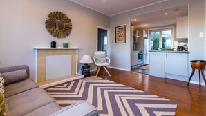 A modern living room with a sofa, a decorative mirror, and a rug. A kitchen is visible in the background with cabinets and a window. - Sunny, Character 2 Bedroom Home - Mission Bay (Auckland)