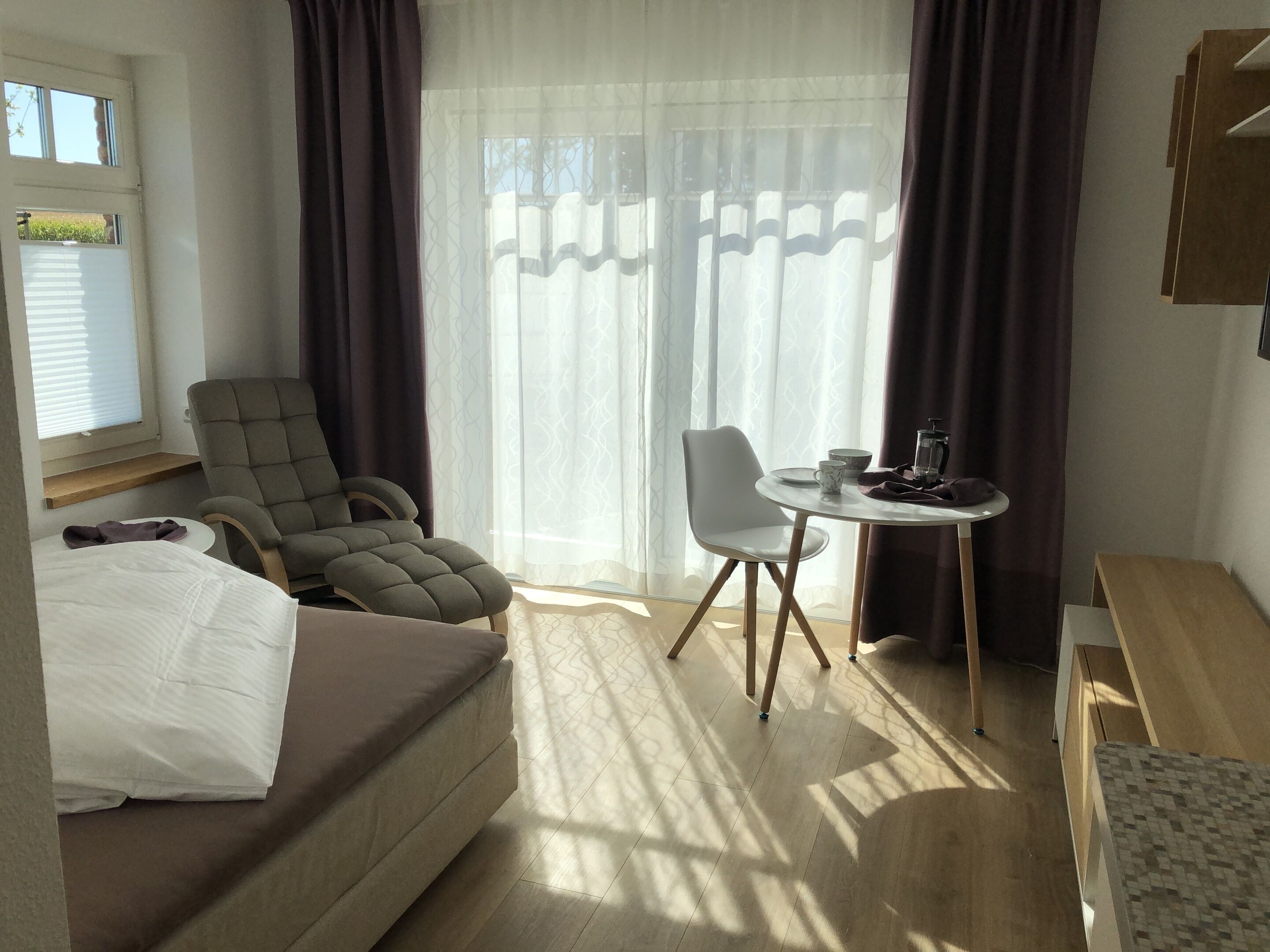 A hotel room with a bed, a grey armchair, a white chair, a small round table with a coffee pot, and a window with curtains.