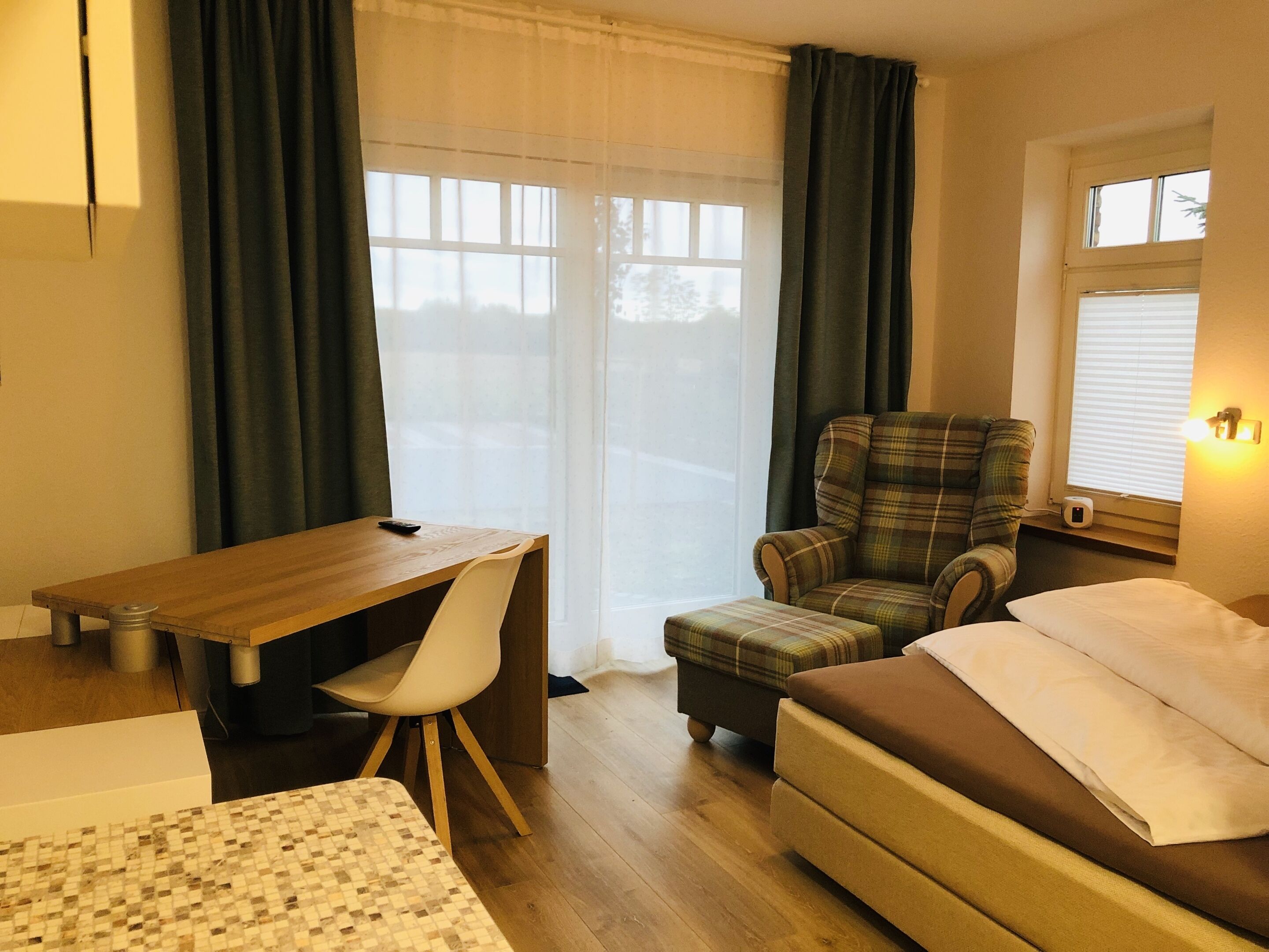 A hotel room with a wooden desk, a plaid armchair, a sofa, and a window with curtains.