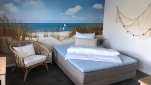 Une chambre avec une fresque murale représentant une plage, un fauteuil en osier, un lit et une décoration en tissu suspendue.