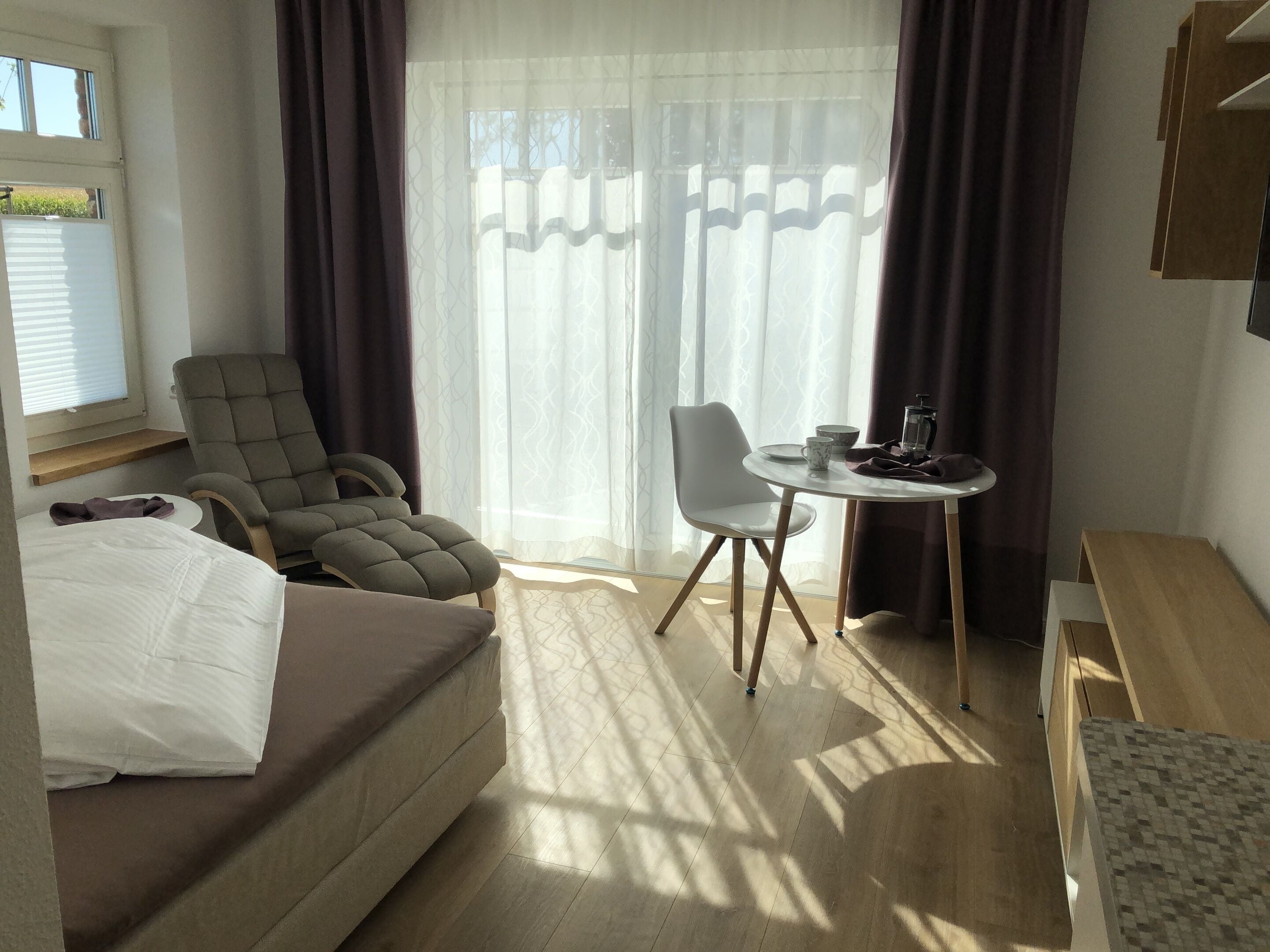 A hotel room with a bed, a sofa, a dining table with chairs, a window with curtains, and a small table with a coffee maker.