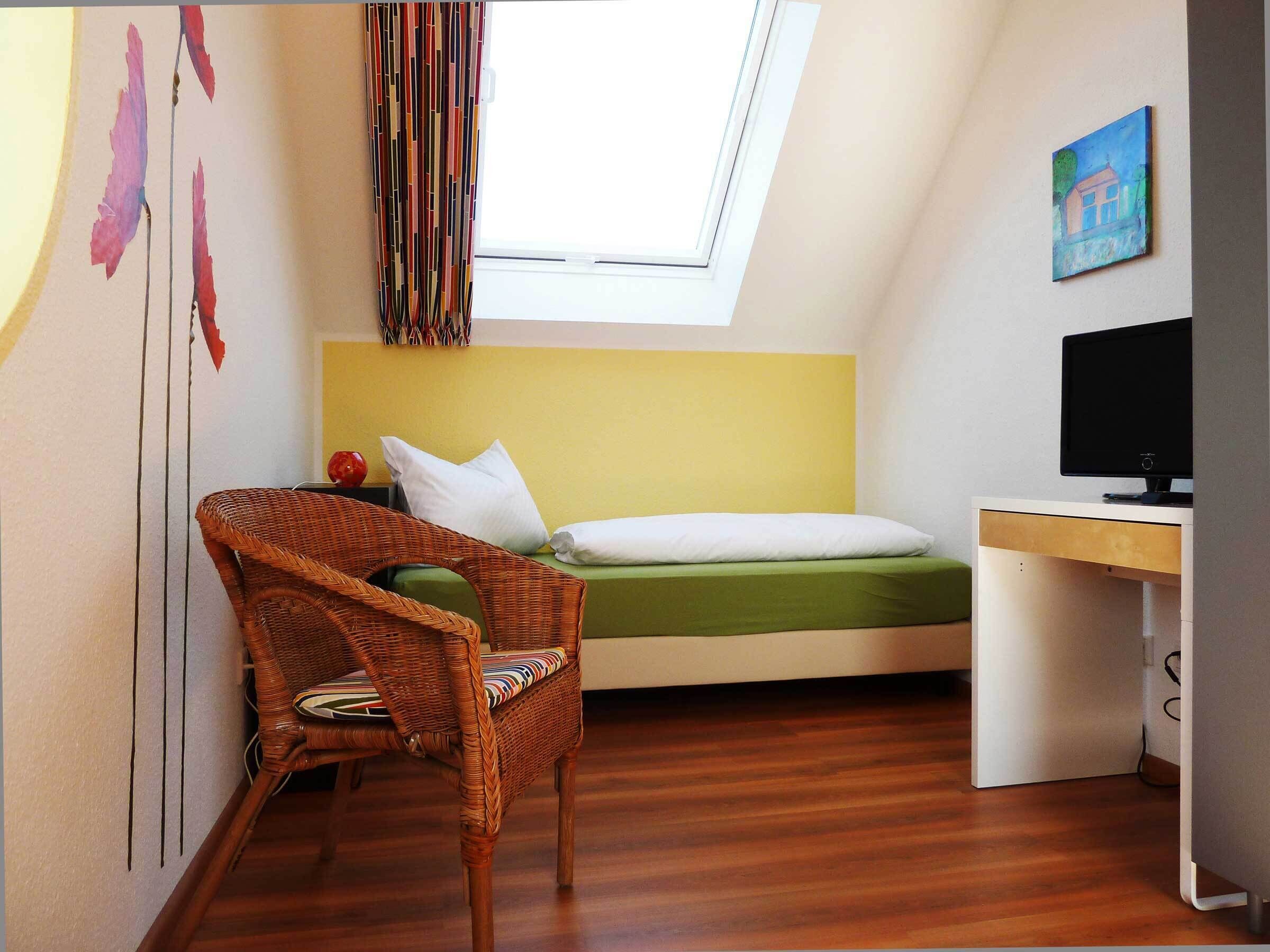 A small, cozy room with a bed, a desk, a wicker chair, a window, and a painting on the wall.