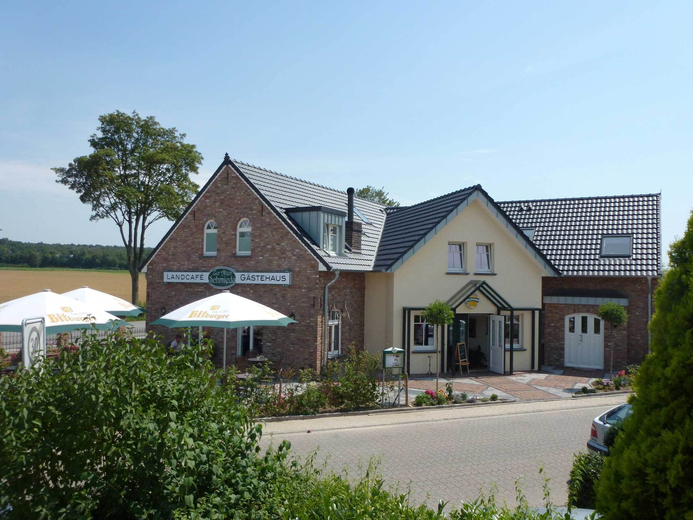 Un edificio de dos pisos con un letrero que dice 'Landcafé Zur Illen Schmidde' y 'Gästehaus'. Hay sombrillas exteriores con la marca 'Bierburger'.