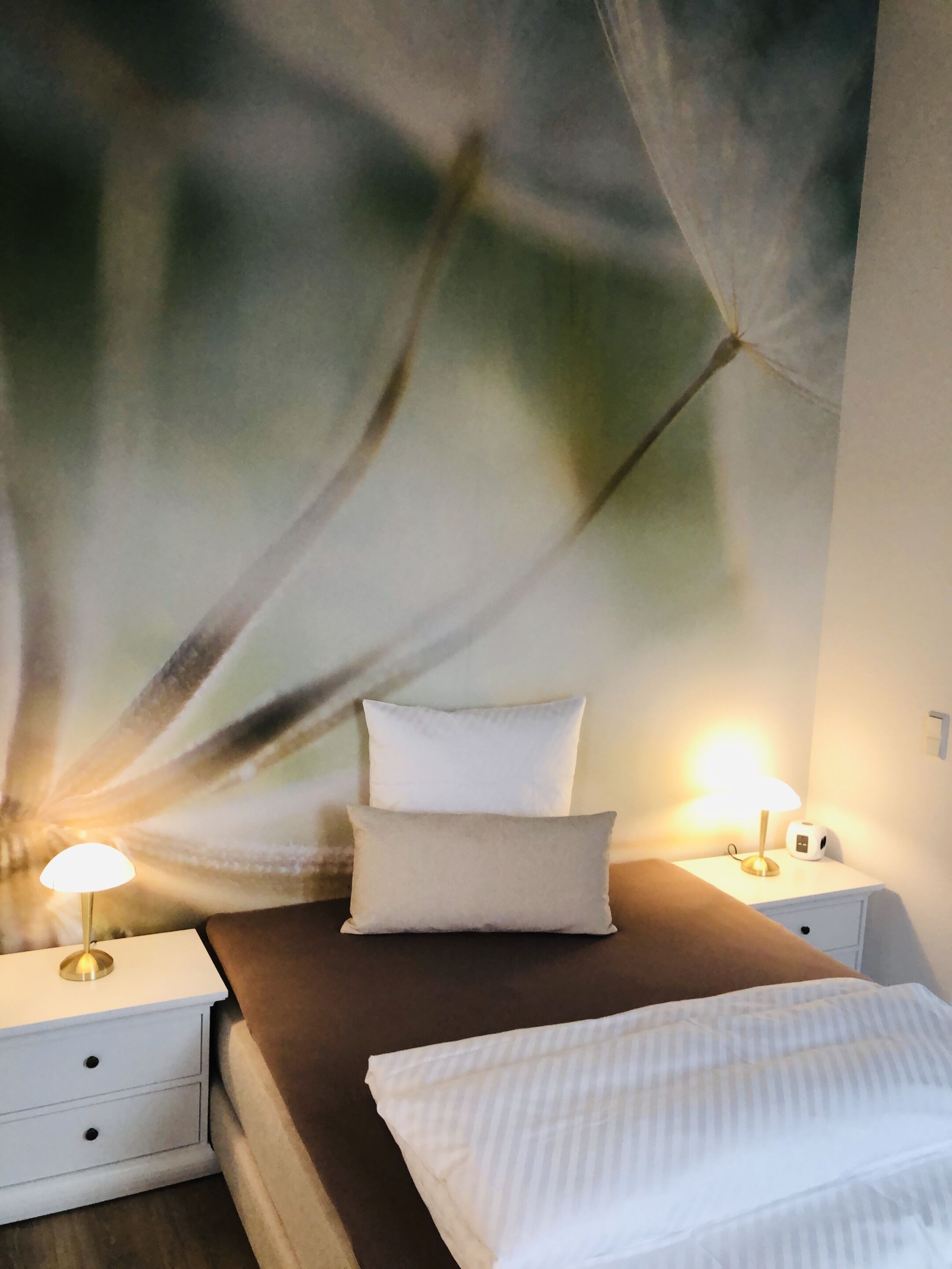 A bedroom with a bed, two bedside tables, a nightstand, a lamp, and a wall with a leaf pattern.