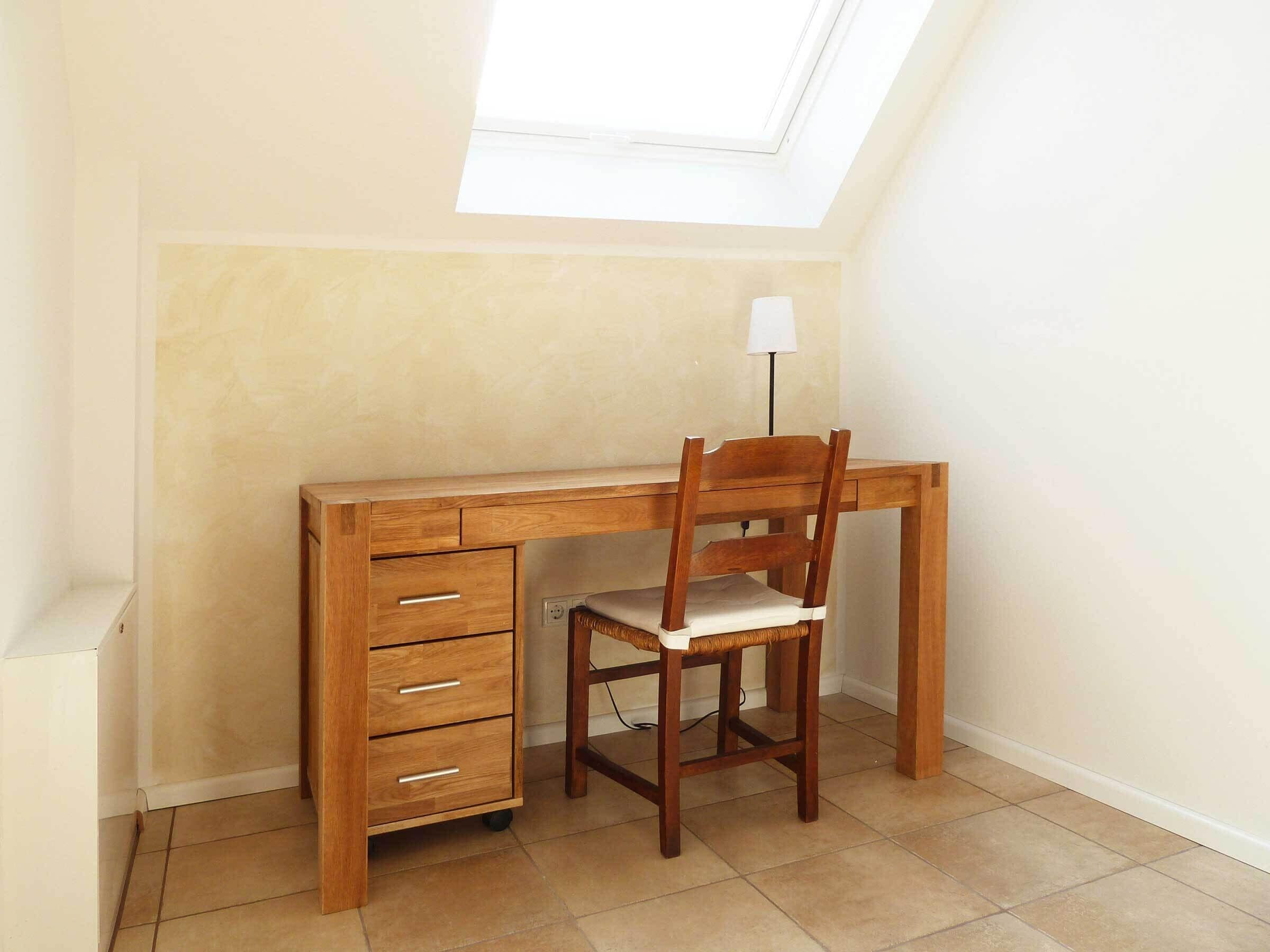 Een houten bureau met een stoel, een lamp en een dakraam in een kamer met betegelde vloer.
