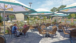 Un espace extérieur aménagé en terrasse, avec des chaises et des tables en osier placées sous des parasols, doté d’un sol en briques et d’un mur en briques en arrière-plan.