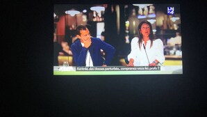 Un écran de télévision diffusant un journal télévisé, montrant deux personnes : l’une porte une veste bleue et l’autre une chemise blanche. Un sous-titre en français apparaît à l’écran.