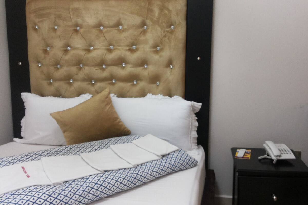 Hotellihuone, jossa on napitettu pääty, kuviollinen vuodevaatteilla varustettu sänky, yöpöytä, jossa on puhelin, ja yöpöytä.