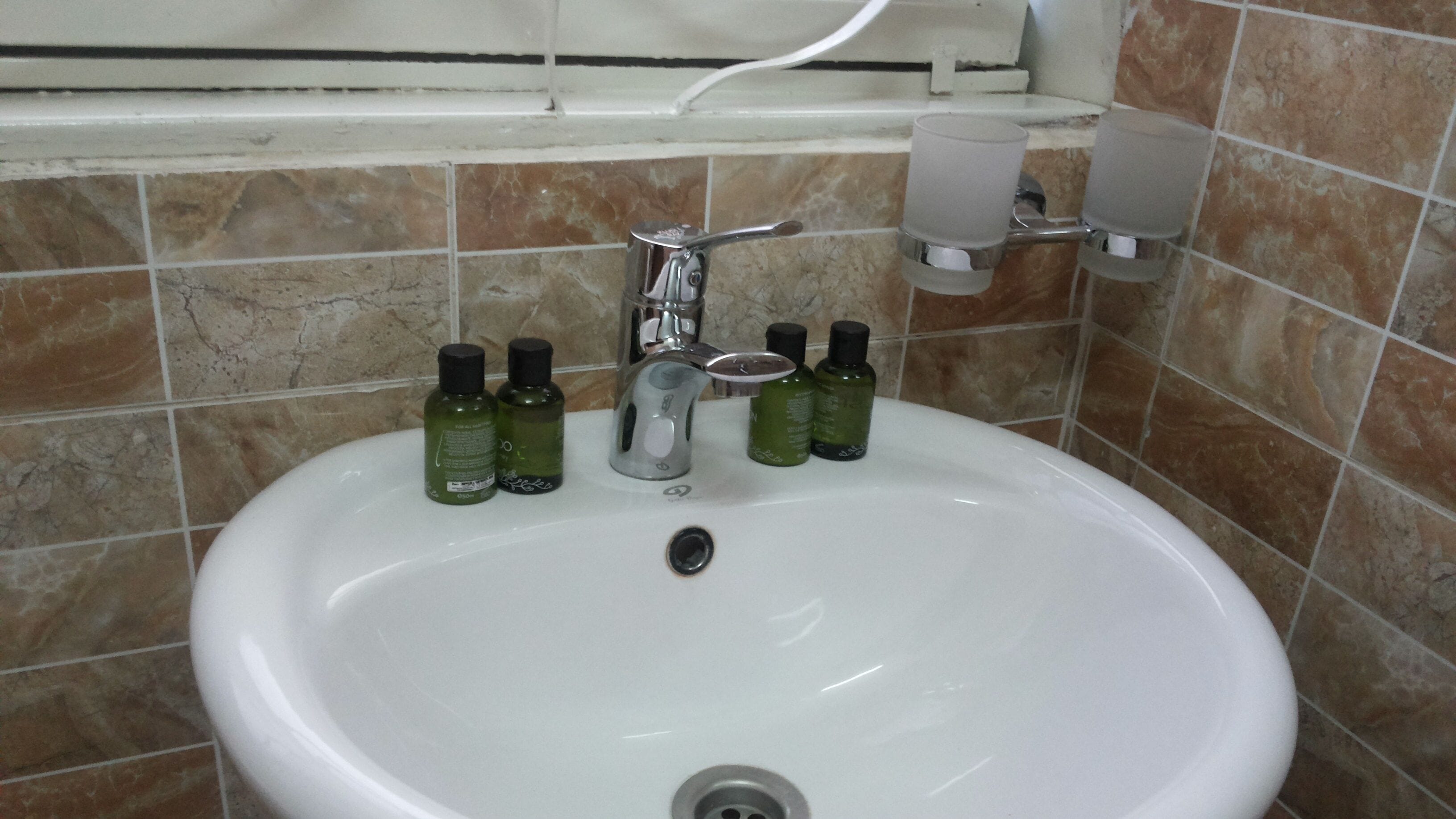 A bathroom sink with a chrome faucet, two green bottles, and a white cup on the wall.