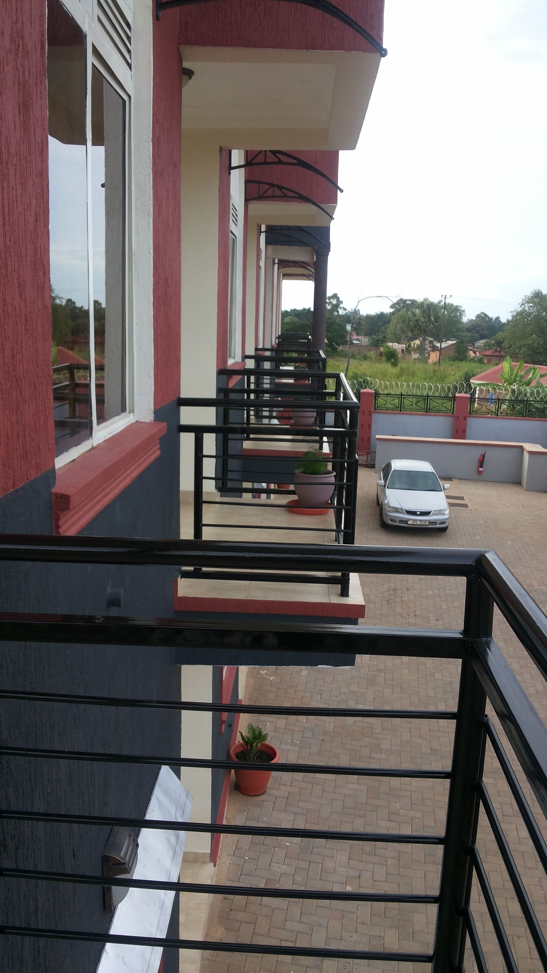 Un bâtiment à la façade rouge et blanche, doté d’un balcon avec une rambarde noire, et d’une zone pavée où se trouvent une voiture garée et des plantes en pot.