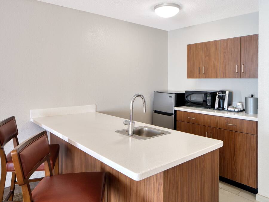 A compact kitchen with a white countertop, stainless steel sink, wooden cabinets, a microwave, and a coffee maker.