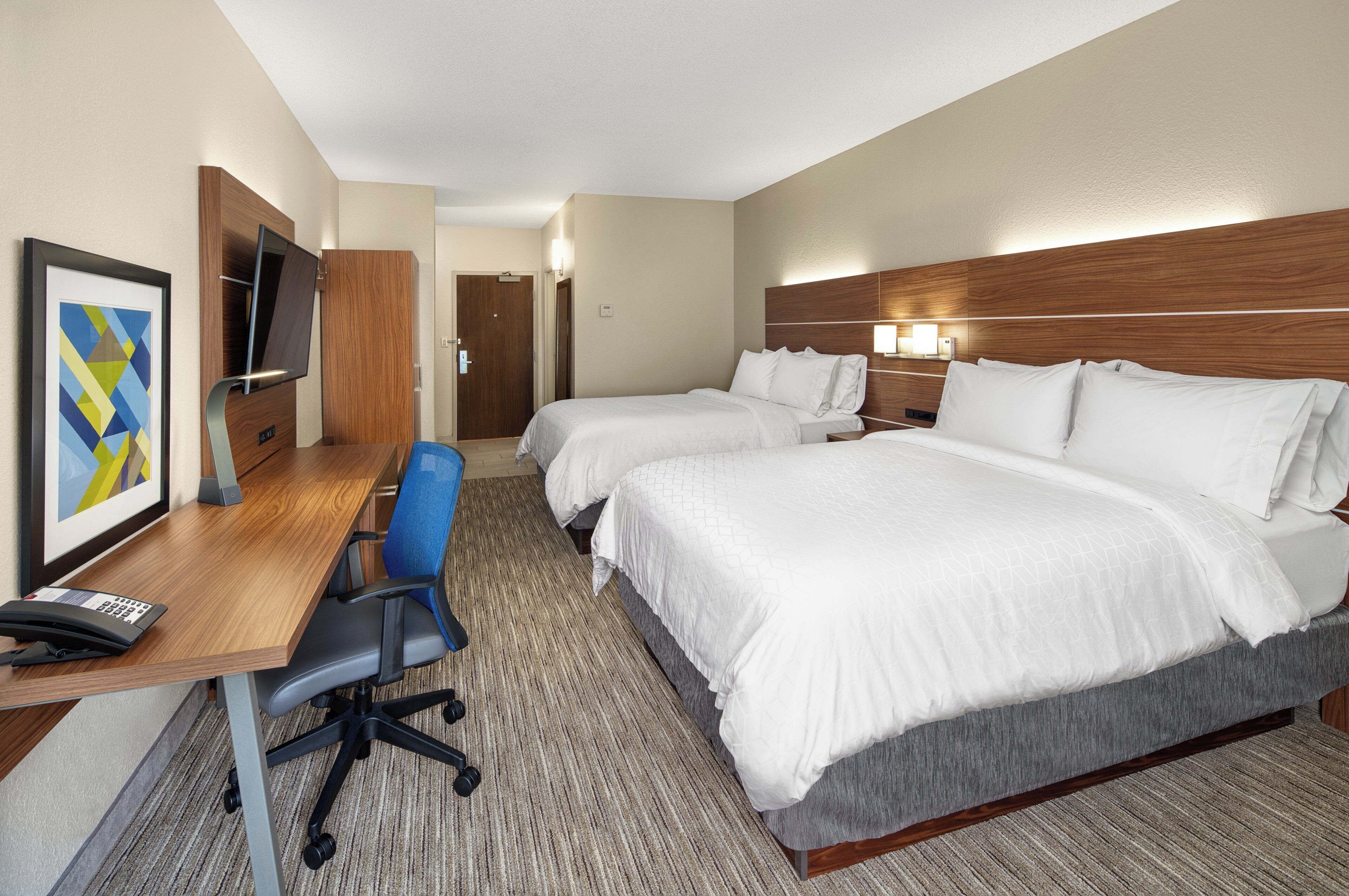 Una habitación de hotel con escritorio, dos camas, una silla, televisión y un cuadro enmarcado.