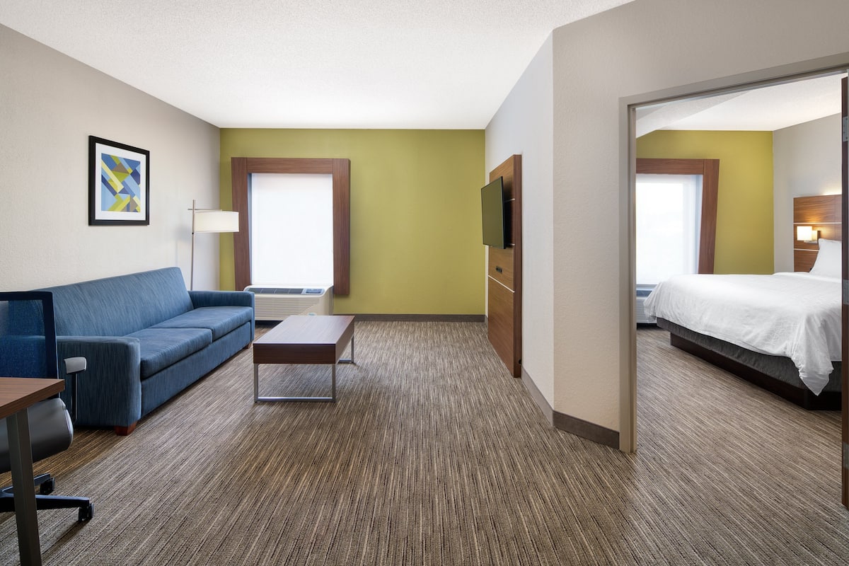 A hotel room with a blue sofa, a small table, a bed, a TV, and a window with blinds.