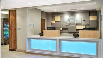Ein Hotel-Empfangsbereich mit modernem Design, inklusive einer Rezeption, eines Automaten und einem Schild, das anzeigt, dass der Shop rund um die Uhr geöffnet ist.