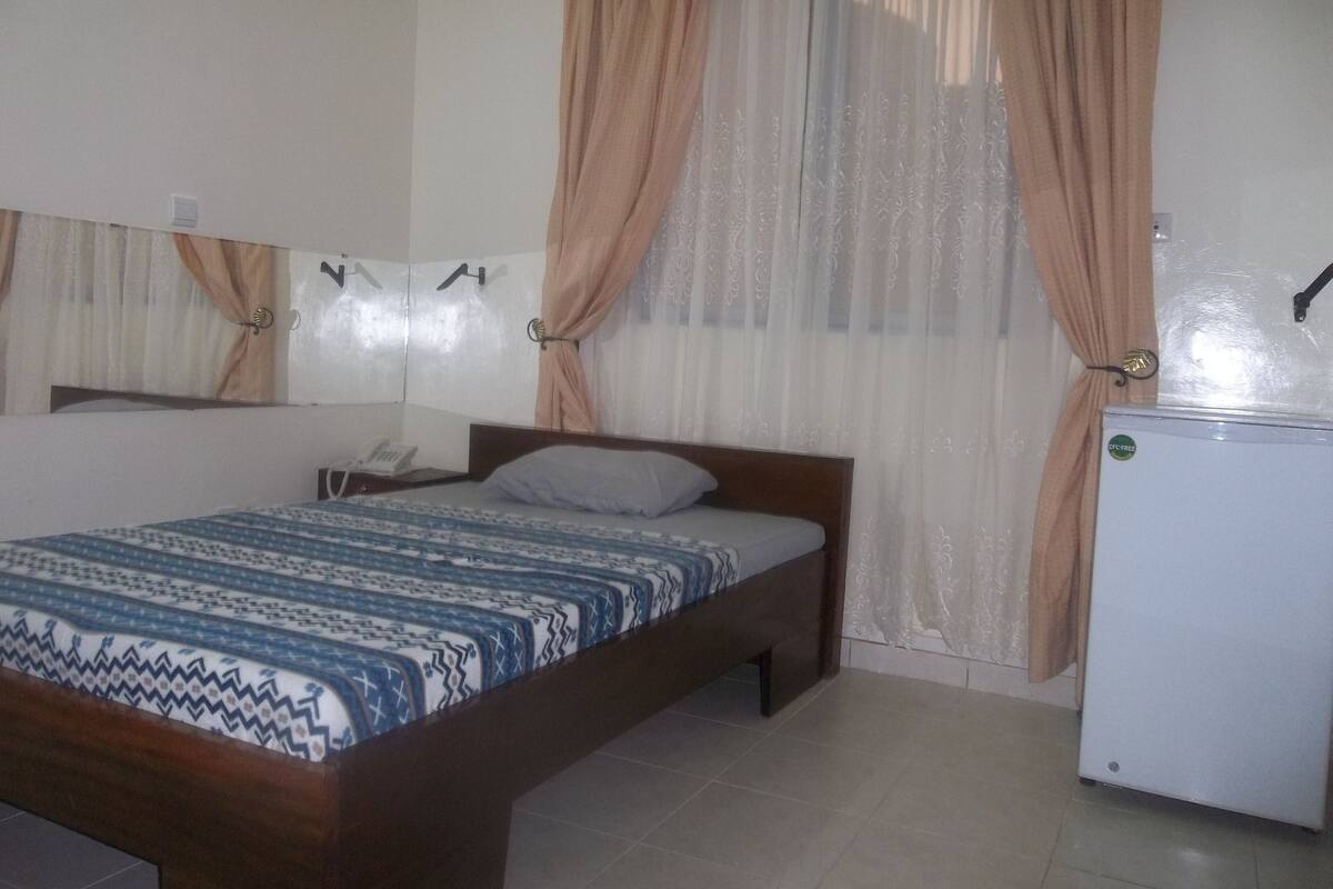 A bedroom with a bed, a bedside table, a telephone, a refrigerator, and curtains.