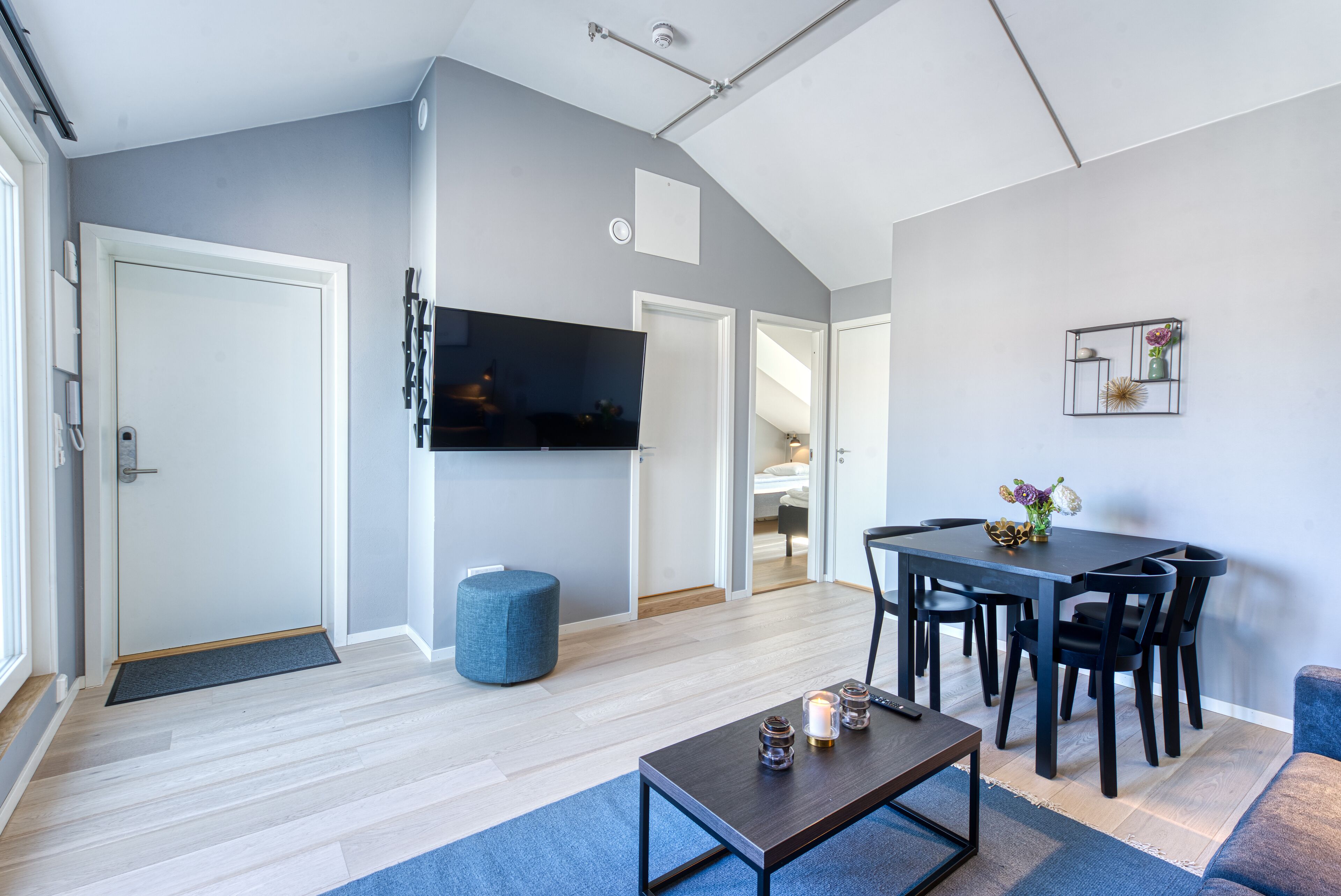 A modern living room with a flat-screen TV, a dining table with chairs, a blue ottoman, and a coffee table.