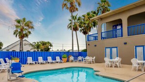A pool area with lounge chairs and tables, palm trees, and a building with blue accents. - Alister Square Inn (Port Aransas)