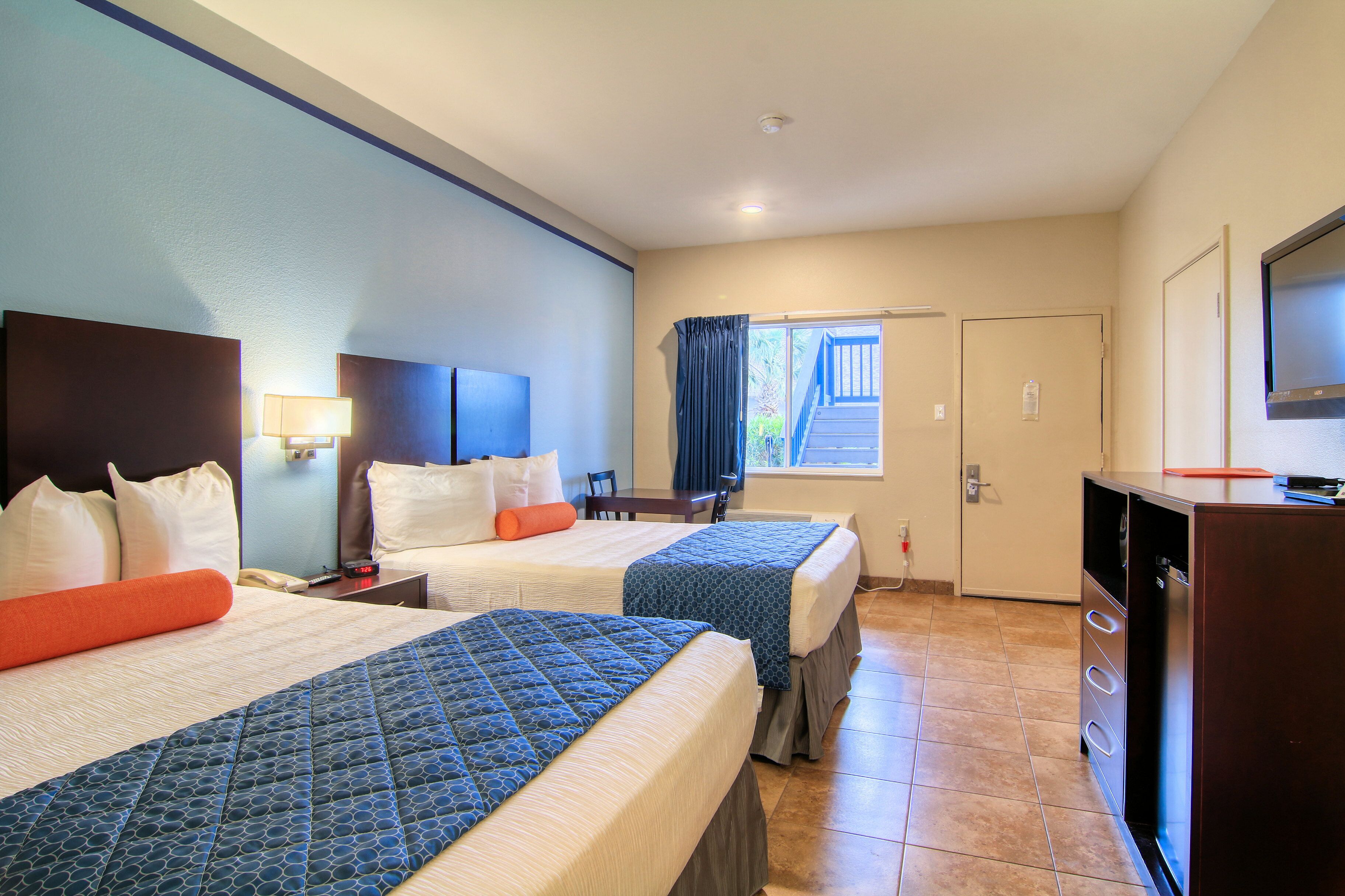 A hotel room with two beds, a desk, a TV, and a window with curtains.