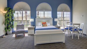 A hotel room with a large bed, a desk, and a chair. There are three arched windows and a large plant. - Alister Square Inn (Port Aransas)