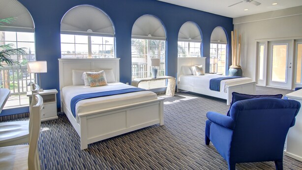 A hotel room with a large bed, a sitting area with a chair, and a view of the outdoors through arched windows. - Alister Square Inn (Port Aransas)