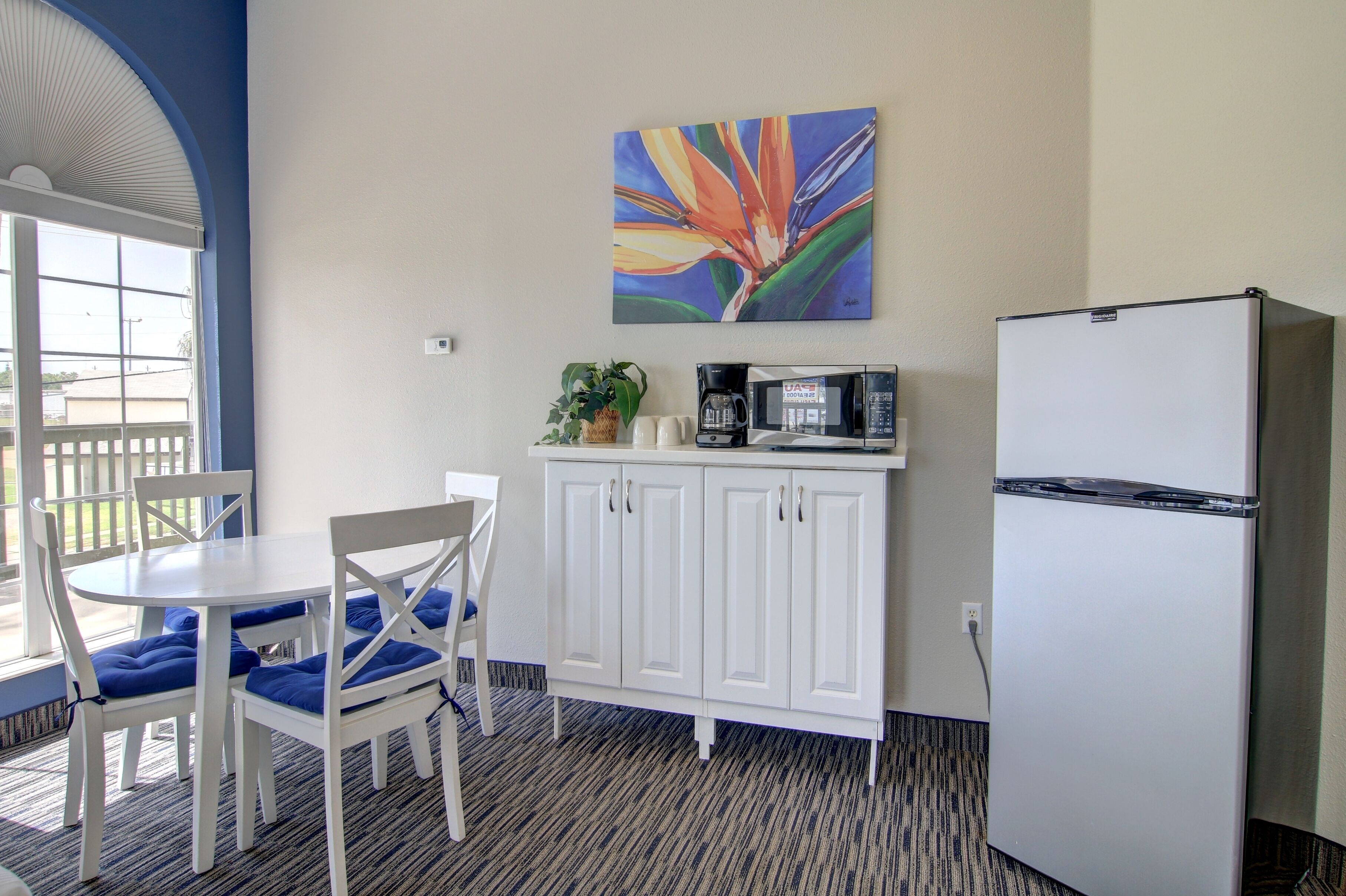 A compact kitchenette with a white refrigerator, a microwave, and a coffee maker on a white cabinet. There is a dining table with blue chairs and a potted plant on the counter.