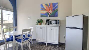 A compact kitchenette with a white refrigerator, a microwave, and a coffee maker on a white cabinet. There is a dining table with blue chairs and a potted plant on the counter.