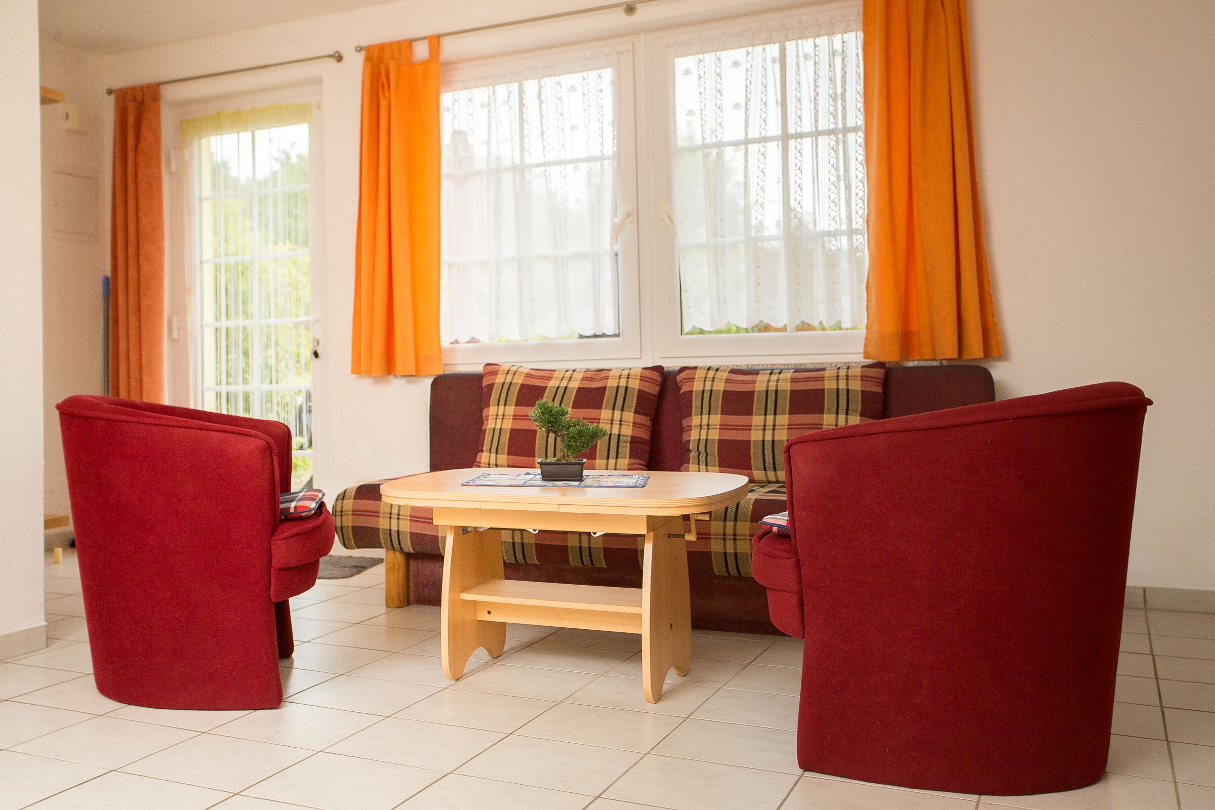 A cozy room with a sofa, two red chairs, a small wooden table, and a window with orange curtains.