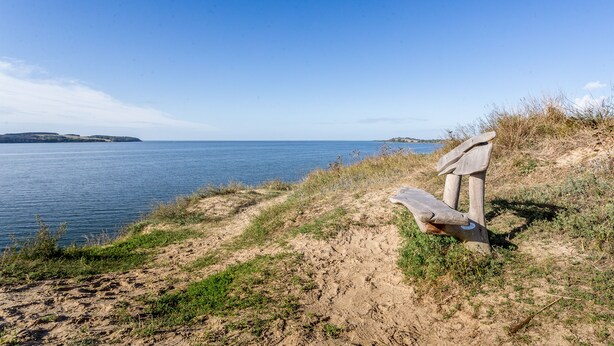 Un banco junto a un sendero costero, con vista al mar y la costa distante.