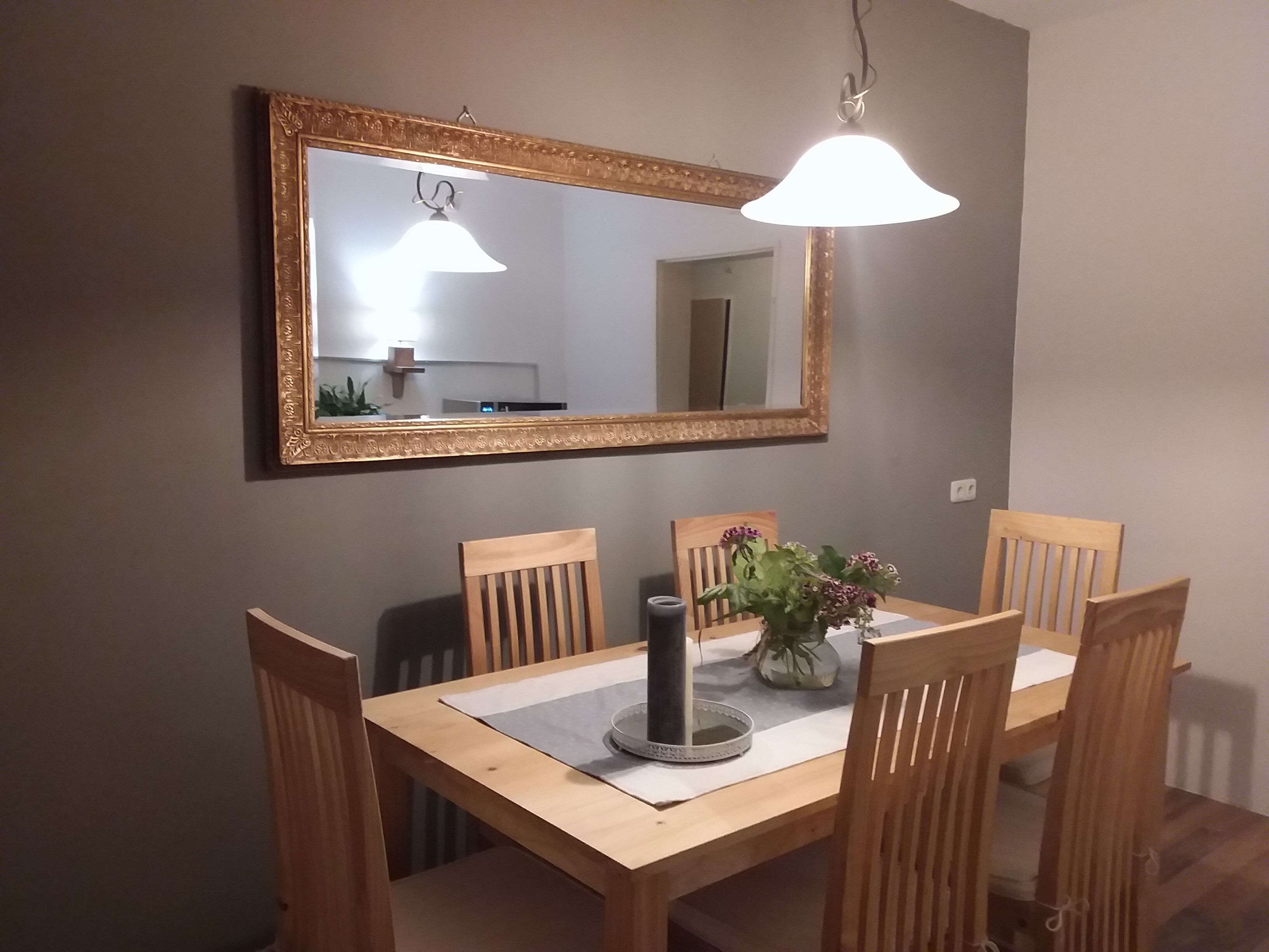 A dining area with a wooden table, four chairs, a vase with flowers, a candle, and a mirror with a gold frame.