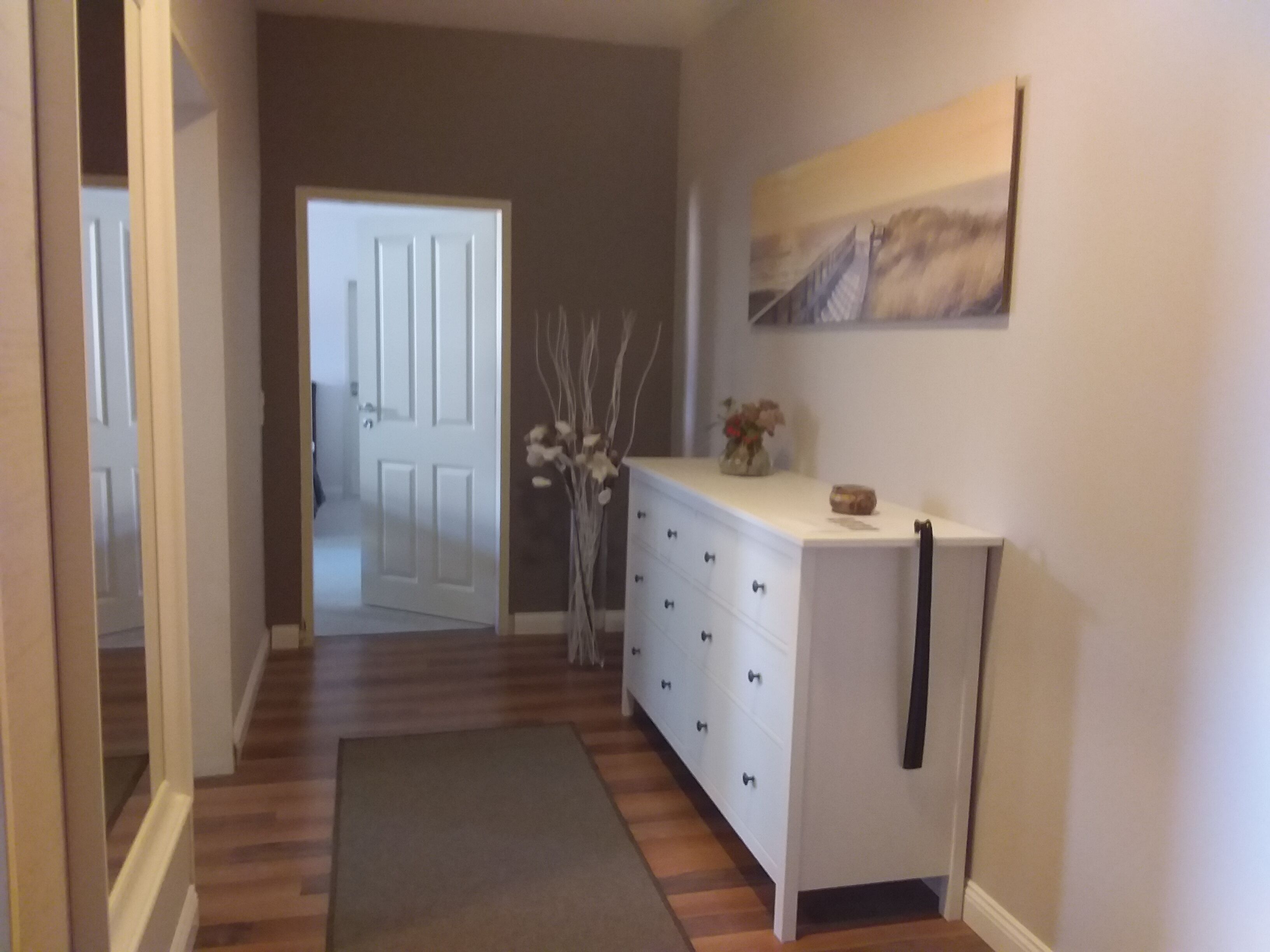 A hallway with a wooden floor, a white dresser with drawers, a vase with flowers, and a door leading to another room.