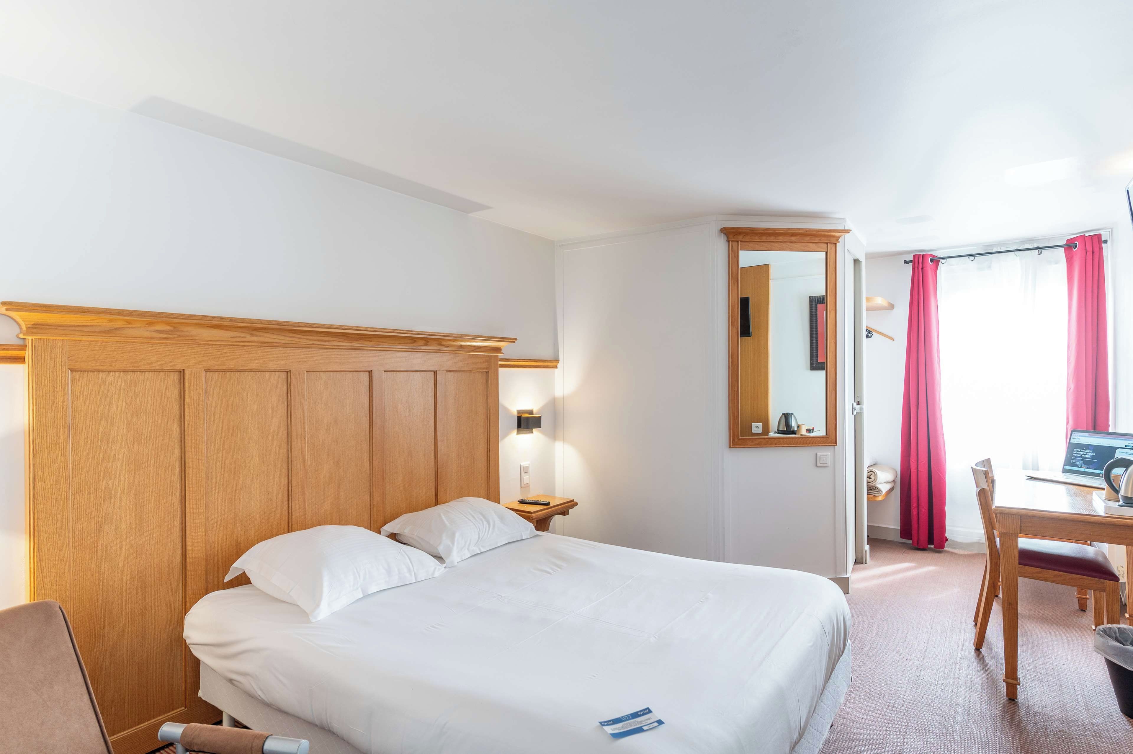 Habitación de hotel con una cama de cabecera de madera, una mesita de noche con lámpara, un escritorio con una computadora portátil y una ventana con cortinas.