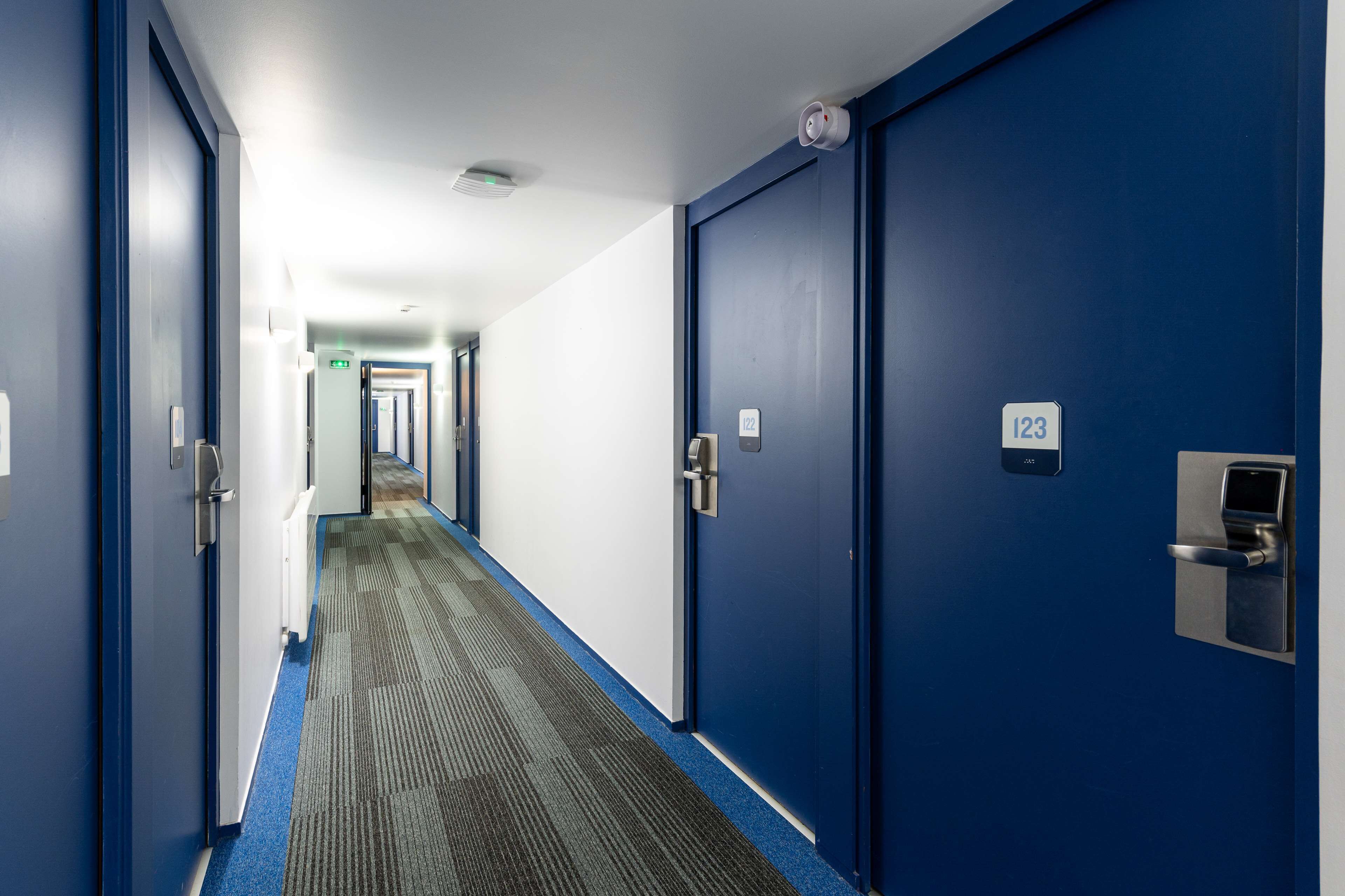 Un couloir d’hôtel avec des portes bleues, des panneaux indiquant le numéro des chambres et un sol recouvert de moquette.