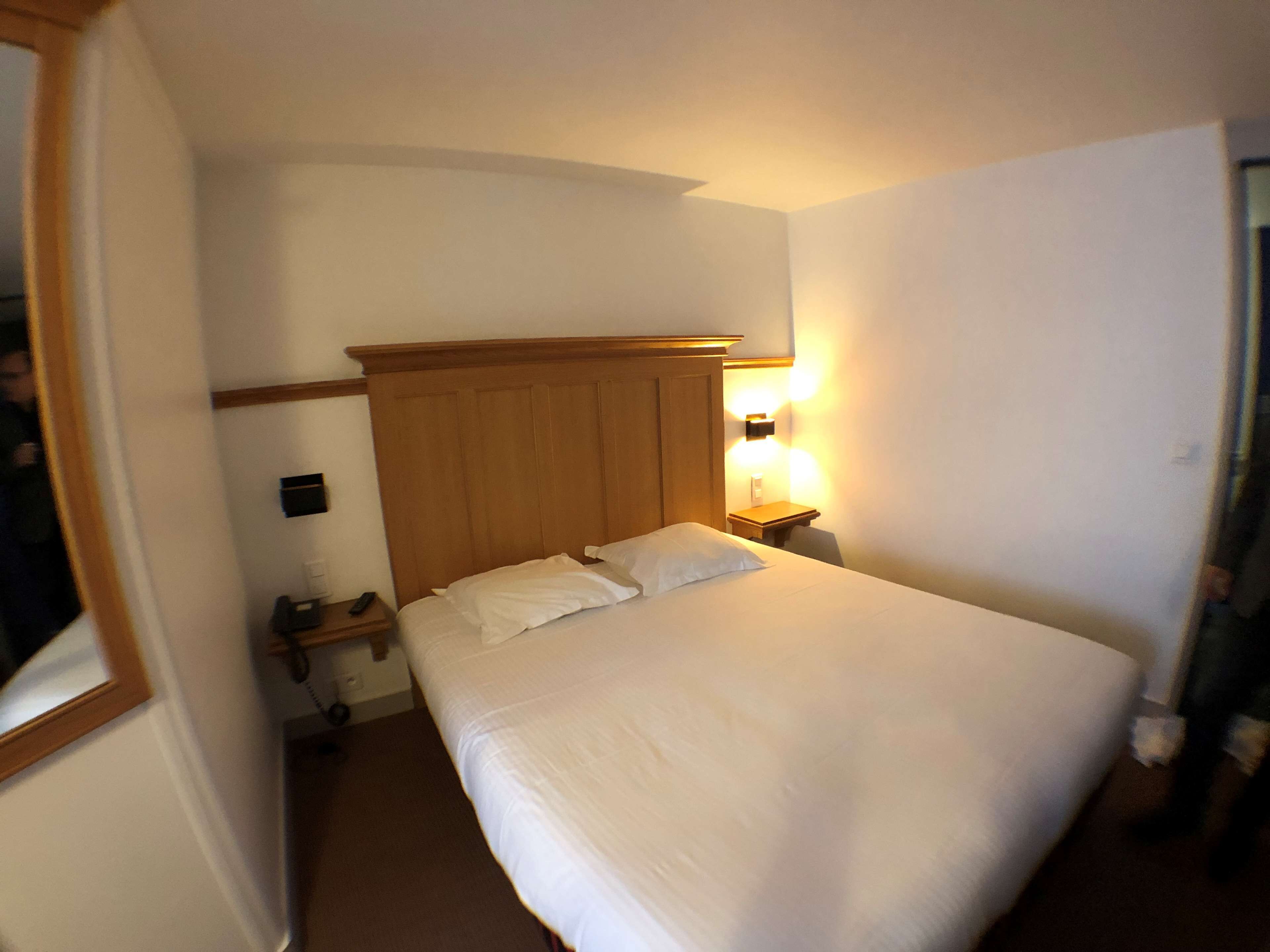 Une chambre d’hôtel avec un lit simple, une tête de lit en bois, une table de chevet avec une lampe et une petite table de nuit avec un téléphone.